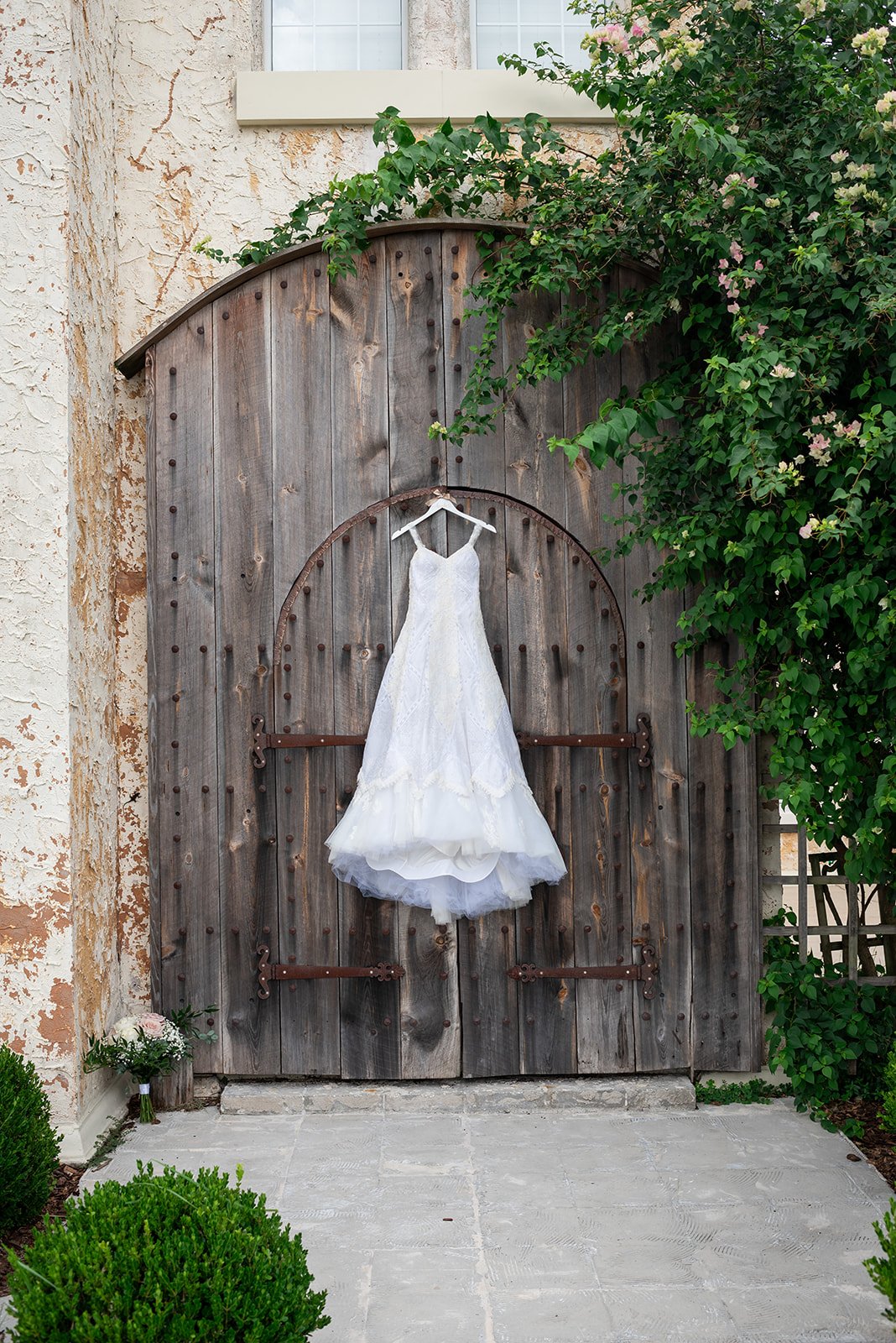 A white wedding dress hanging on a rustic wooden door, surrounded by greenery and flowers.