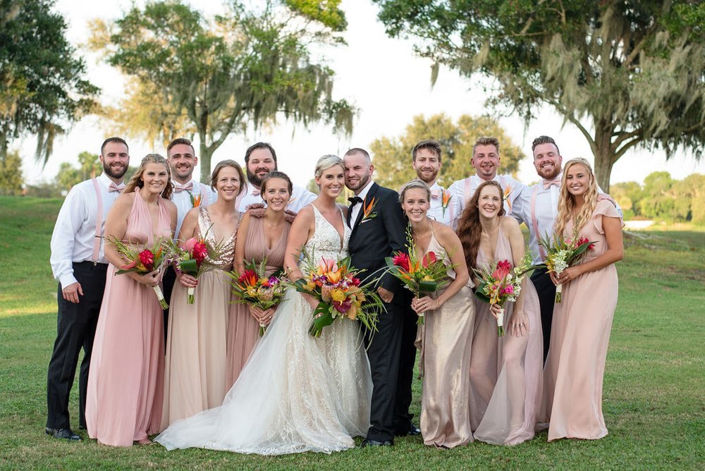 A wedding party posing outdoors, featuring a bride in a white gown and groom in a black suit, surrounded by bridesmaids in pastel dresses holding bouquets, and groomsmen in white shirts with corsages. The background includes trees and grass under a c