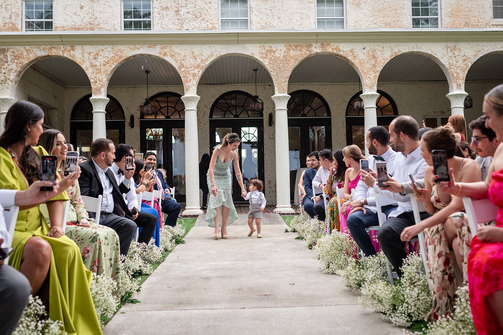 A woman and a young child walk down an outdoor aisle, surrounded by seated people taking photos, during a wedding ceremony with a rustic building in the background.