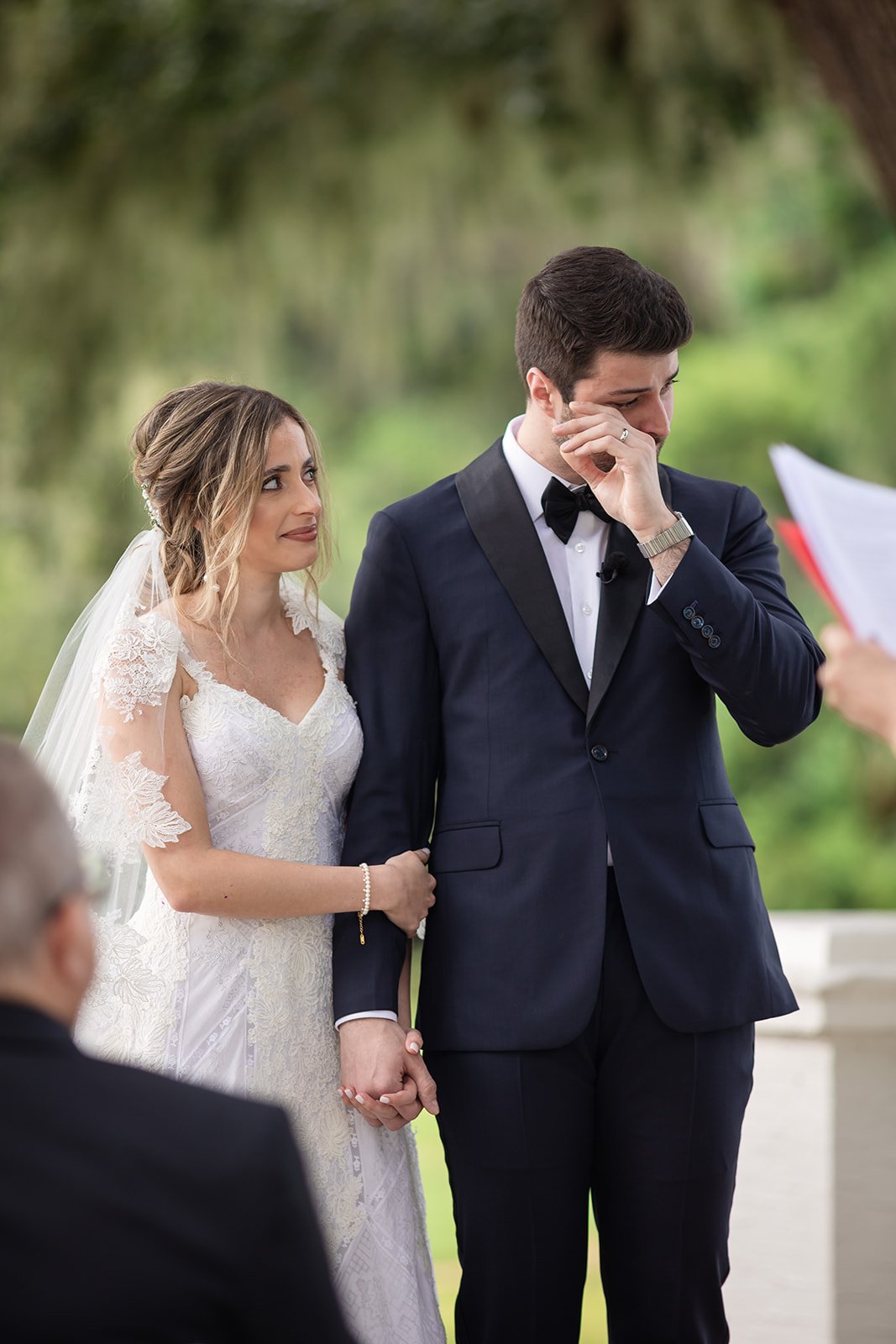Bride and groom holding hands during an outdoor wedding ceremony, with the groom wiping his eye.