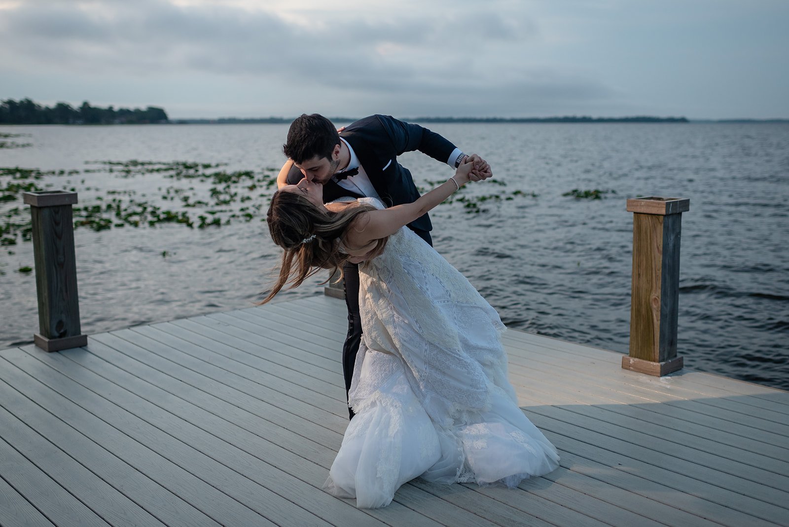 Bride and groom dancing on a wooden pier by the water during sunset.