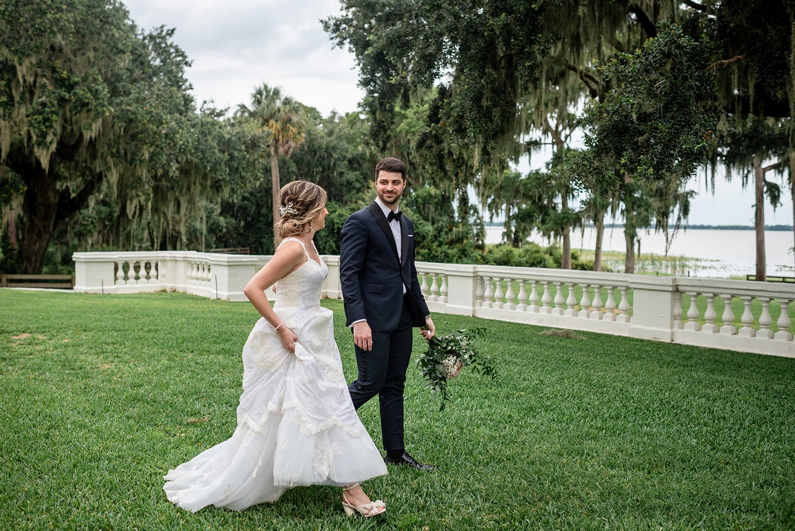 Bride and groom walking on a grassy lawn with a scenic backdrop of trees and a lake.