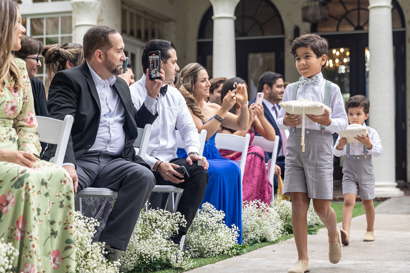 Wedding ceremony with young ring bearers holding pillows walking down the aisle, guests seated and taking photos in the background.