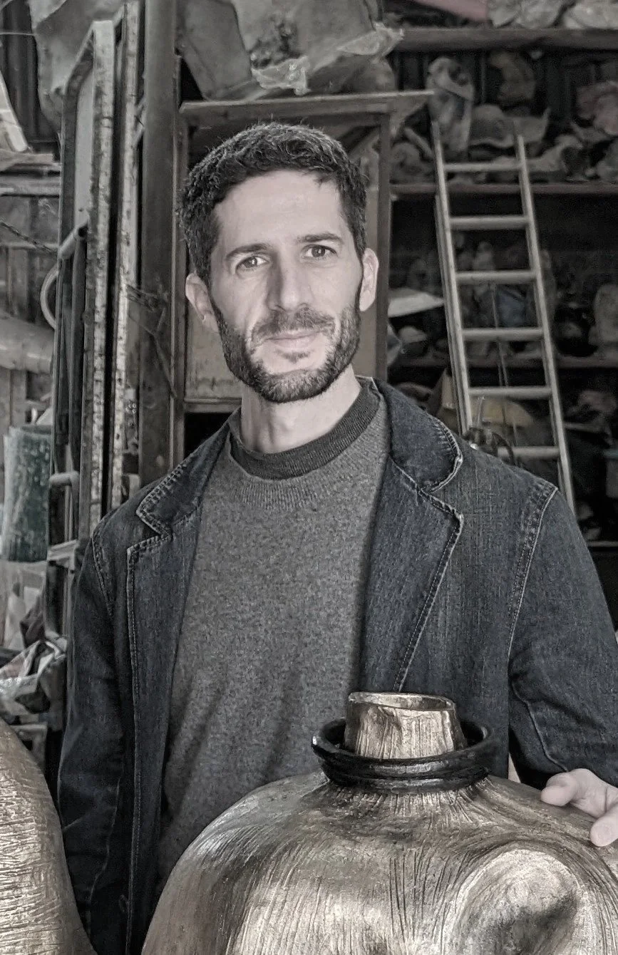 A man with dark, curly hair and a beard standing in a workshop, holding a round, wooden object. He is wearing a dark-colored denim jacket over a gray sweater. The background shows shelves and ladders filled with tools and materials.