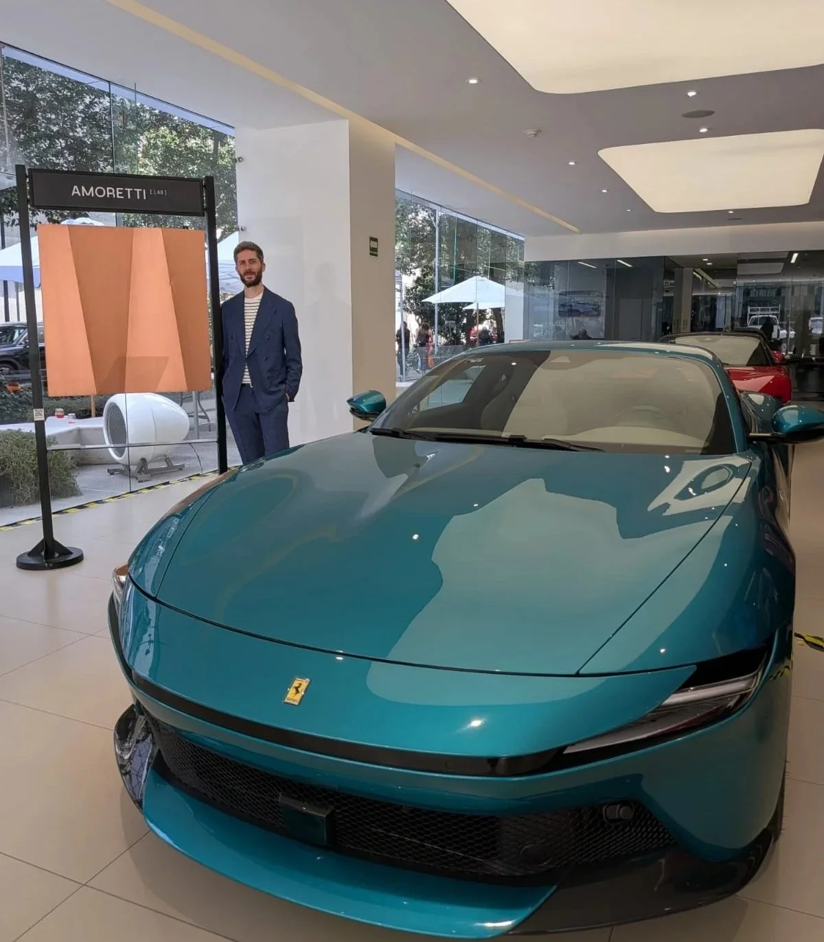 Andrea Amoretti junto a un panel de cobre trabajado a mano de AmorettiLAB y un Ferrari turquesa en el showroom de Ferrari, Ciudad de México