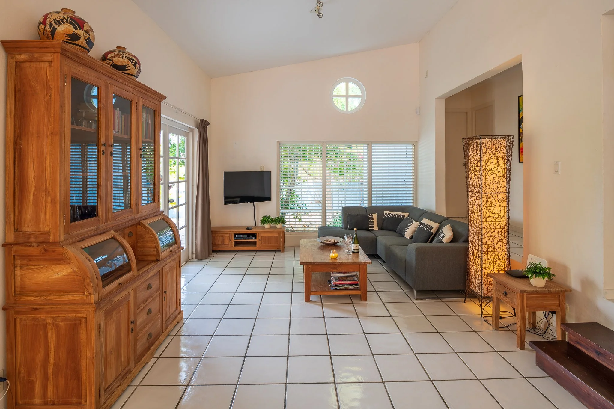 Living room with tiled floors, a gray sectional sofa with black and white cushions, a wooden coffee table with magazines, a TV on a wooden stand, a tall wicker floor lamp, a wooden display cabinet with glass doors, a small side table with a plant, a 