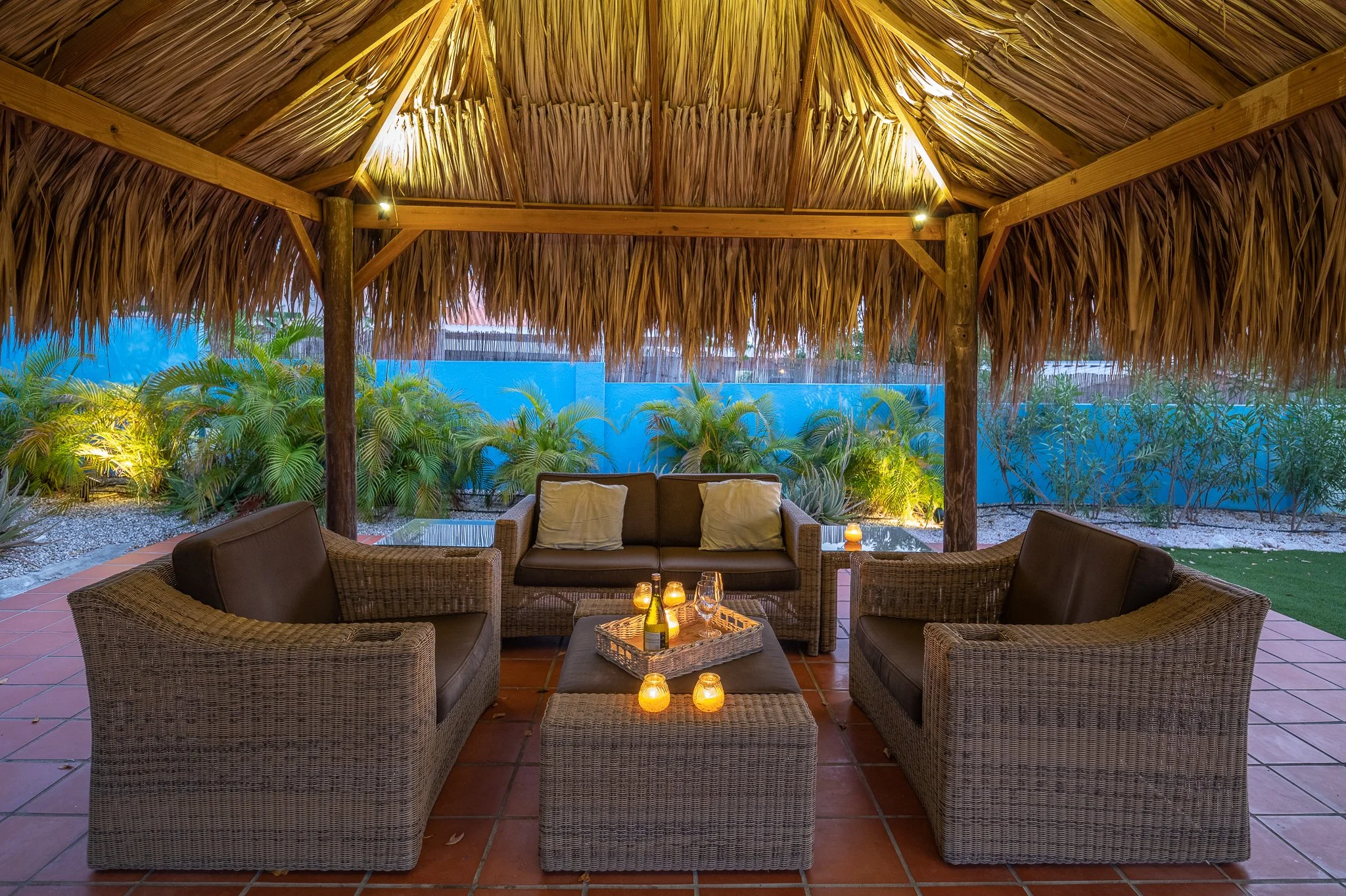 Outdoor lounge area with wicker sofas and a thatched roof, illuminated by candles and soft lighting, with tropical plants in the background.