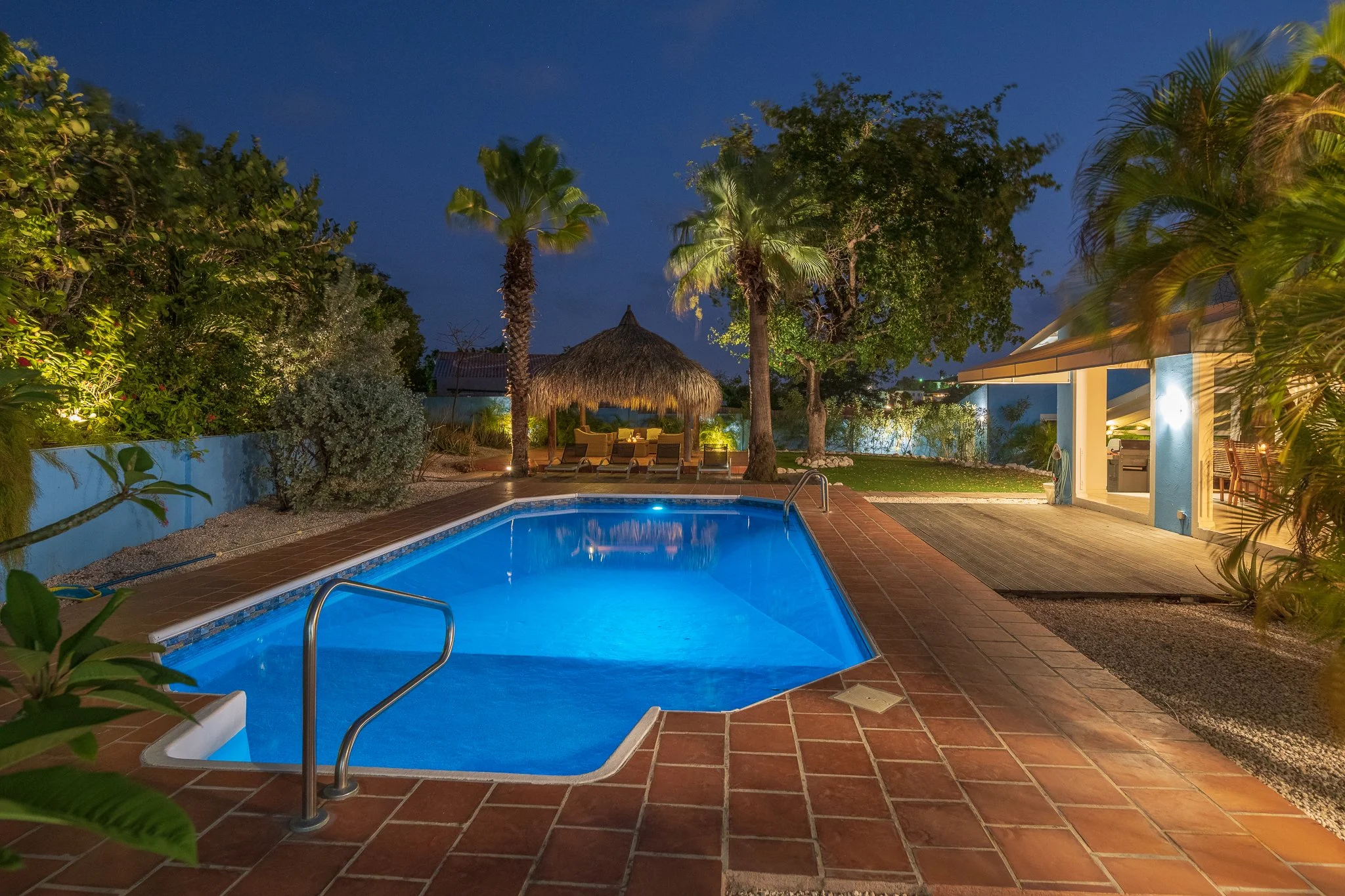 Night view of a backyard swimming pool illuminated with blue lights, surrounded by tropical plants, trees, and a thatched hut with lounge chairs underneath. There is a poolside building with an open area and a wood deck.