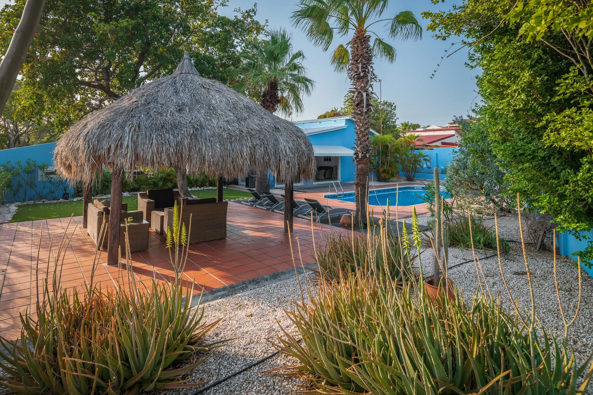 A backyard patio with a thatched umbrella, outdoor sofa, and tables near a swimming pool, surrounded by green plants and trees.