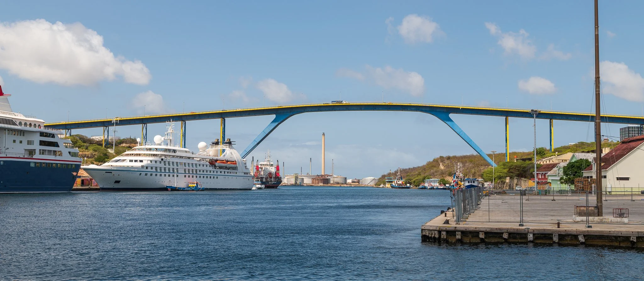 View of a harbor with several large white yachts docked, a blue and yellow bridge overhead, and a hillside with houses in the background under a partly cloudy sky.