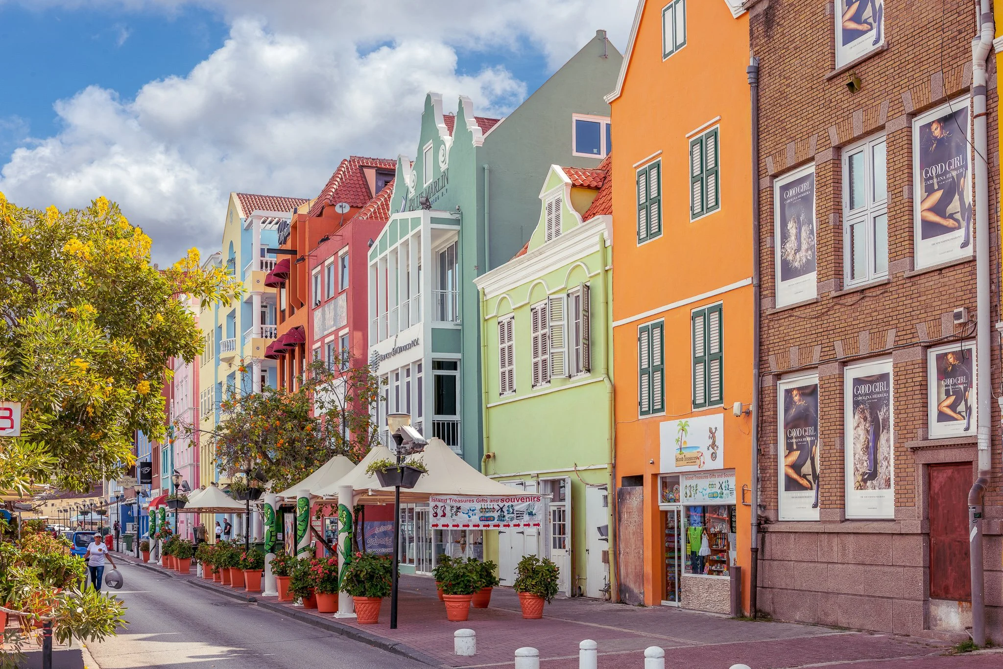 Colorful buildings along a street with outdoor umbrellas, potted plants, and a lamppost, under a partly cloudy sky.