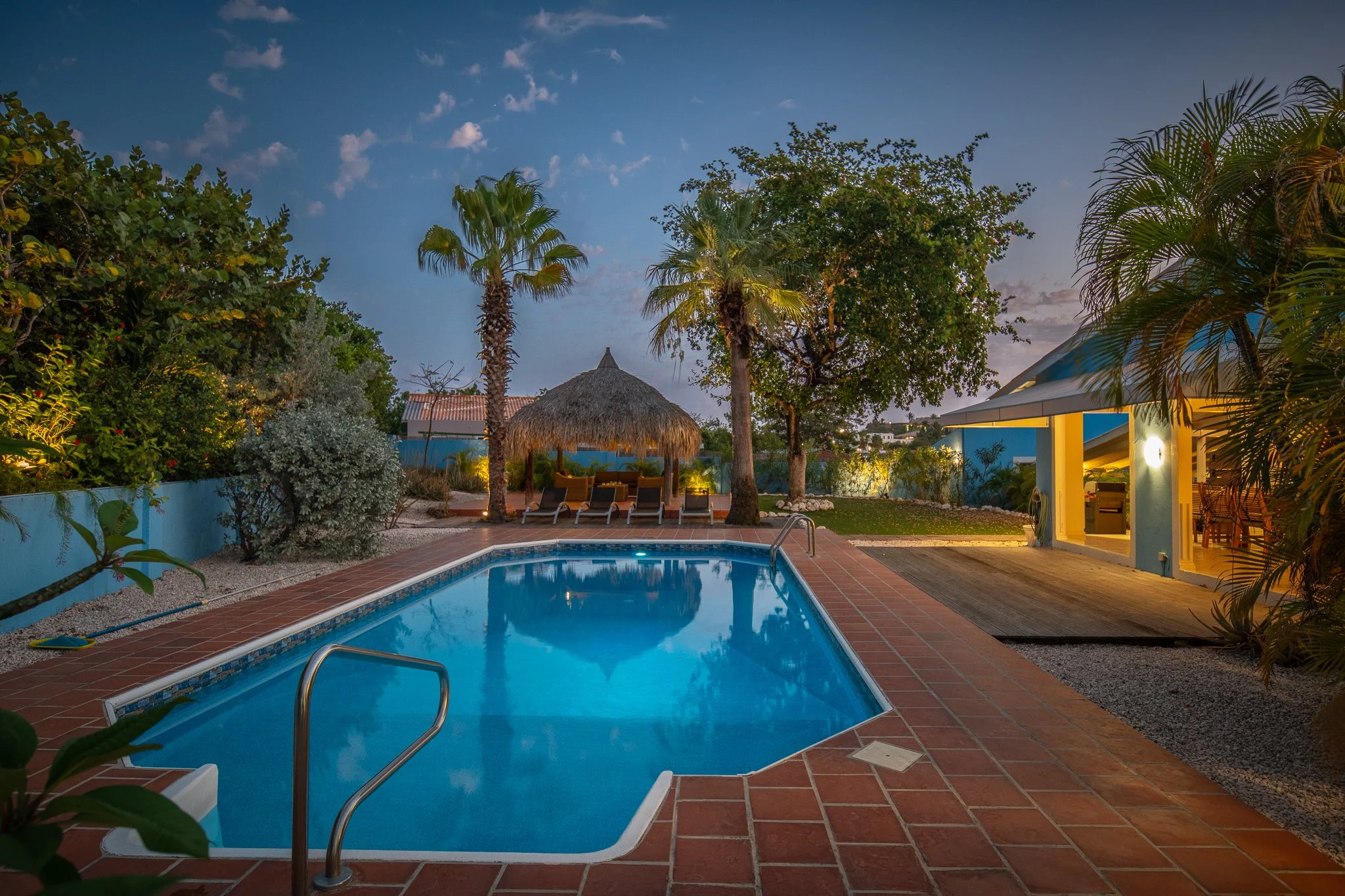 A backyard pool area at dusk with a blue pool, surrounded by tropical plants, with a thatched hut, lounge chairs, and a pool house illuminated warmly.