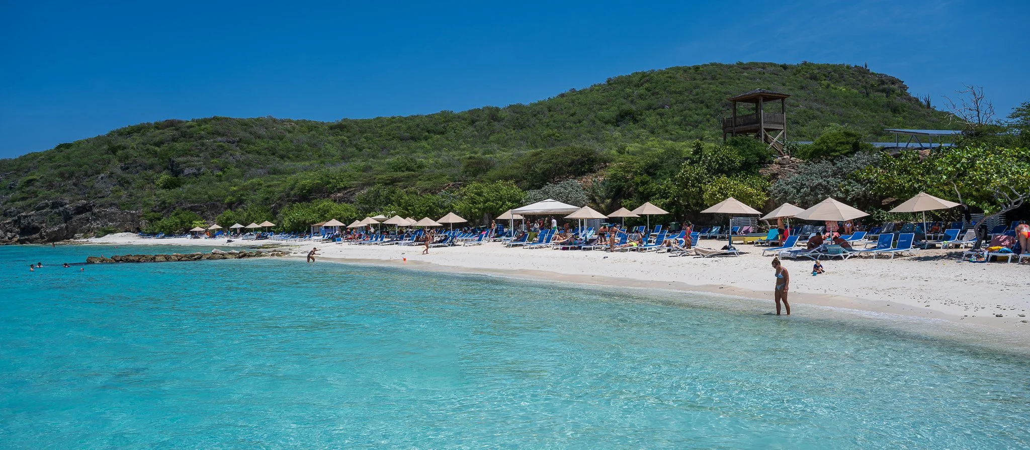 A beach with clear turquoise water, white sand, and sun loungers under umbrellas. Hills covered with green vegetation are in the background, along with a wooden lookout tower and a small structure. People are relaxing, swimming, and walking along the shoreline.