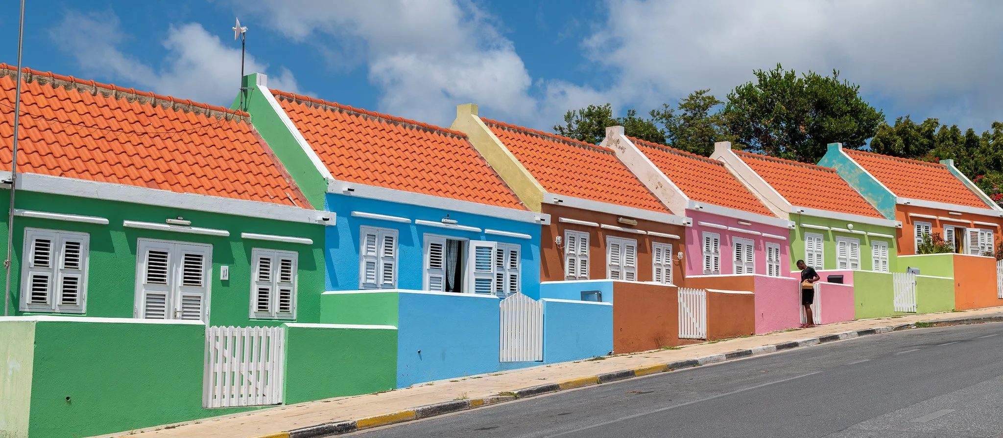 Colorful row of houses with painted walls in green, blue, brown, pink, lime green, and orange with orange tiled roofs under a partly cloudy sky. One person stands outside a house near the pink house.