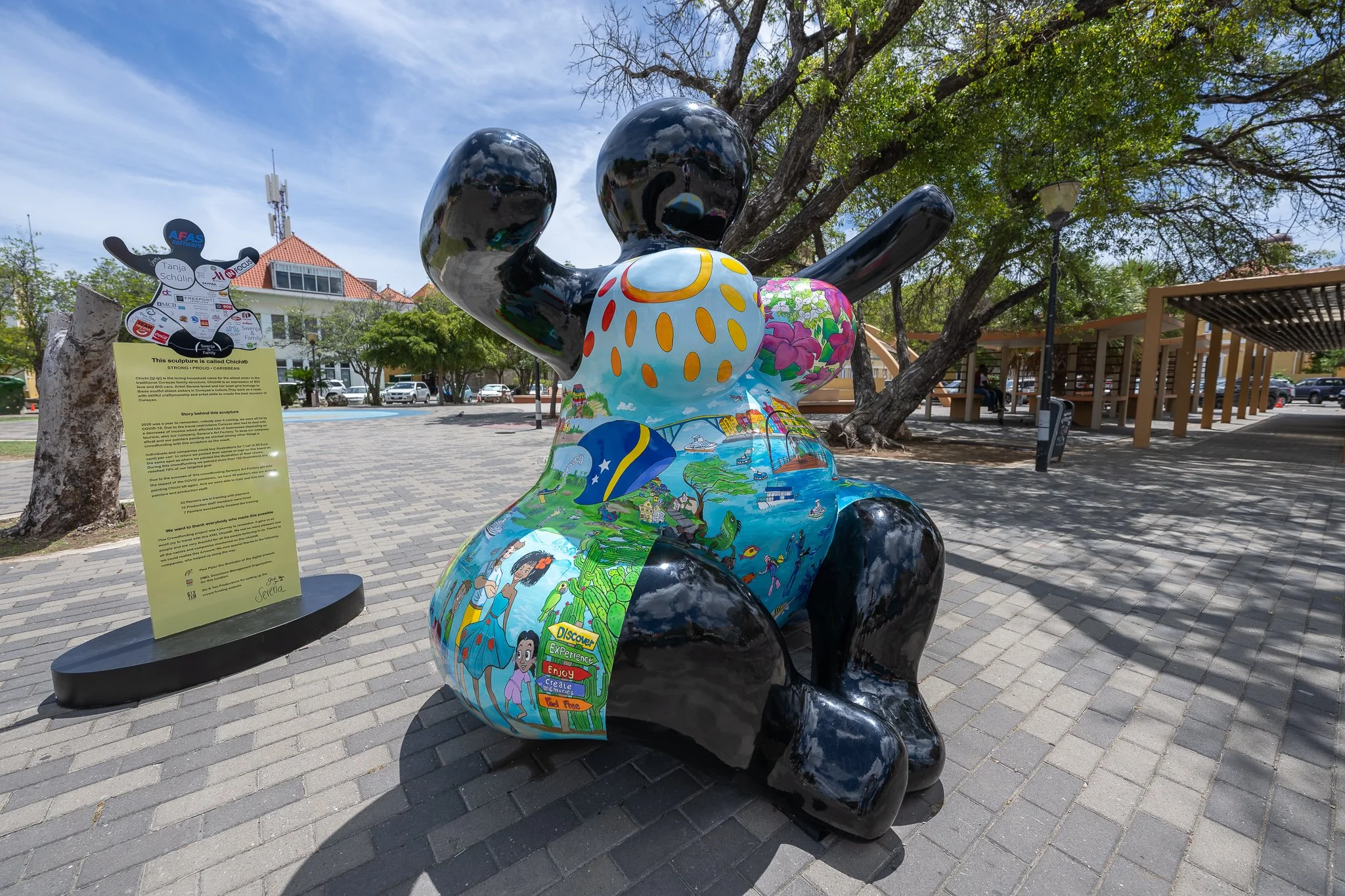 Colorful sculpture called 'Chichi' in a park with trees, a sign, and benches.