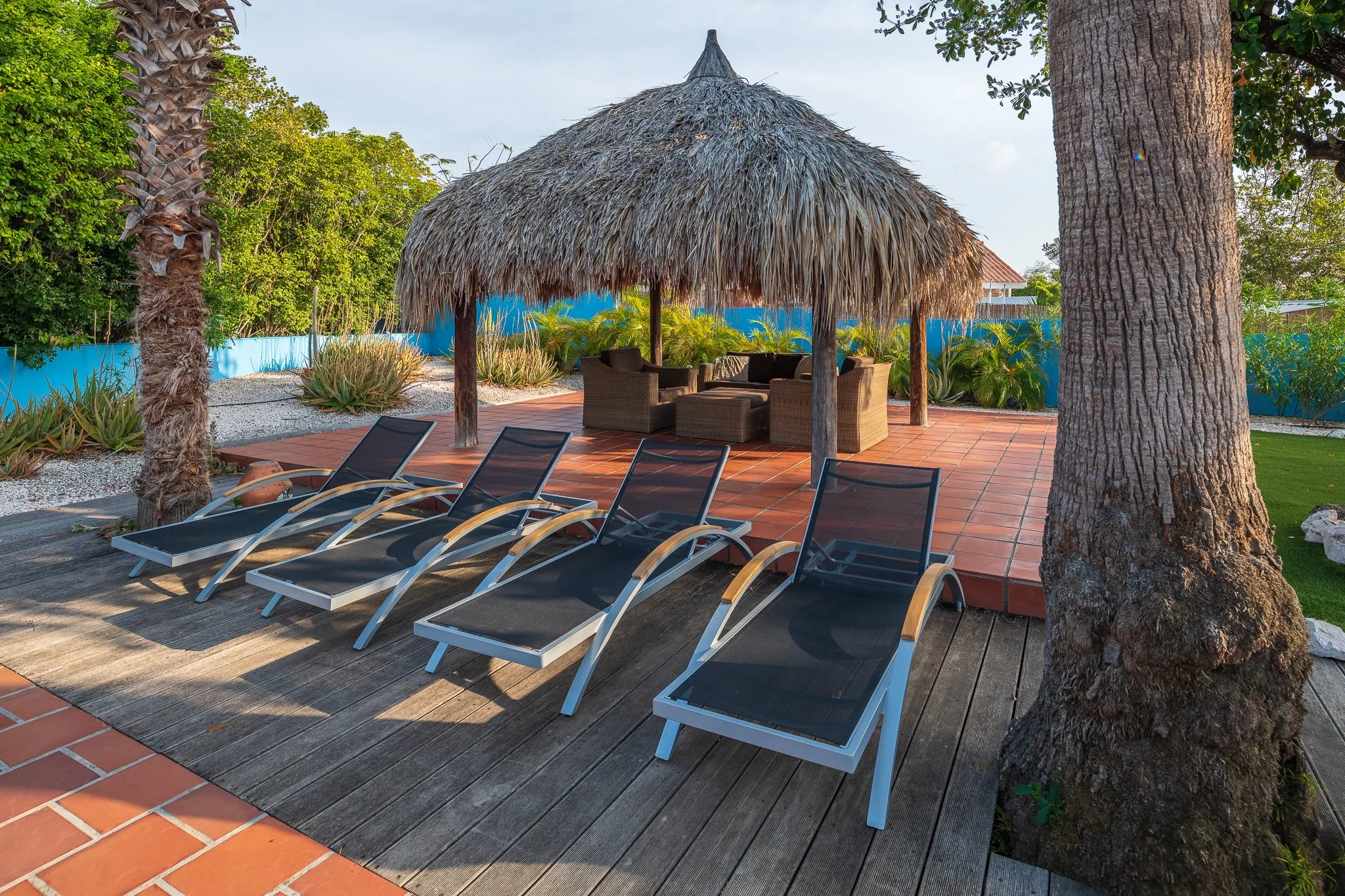 A tropical outdoor lounge area with four black lounge chairs, a thatched roof hut with wicker furniture, palm trees, and desert plants on a tiled patio.