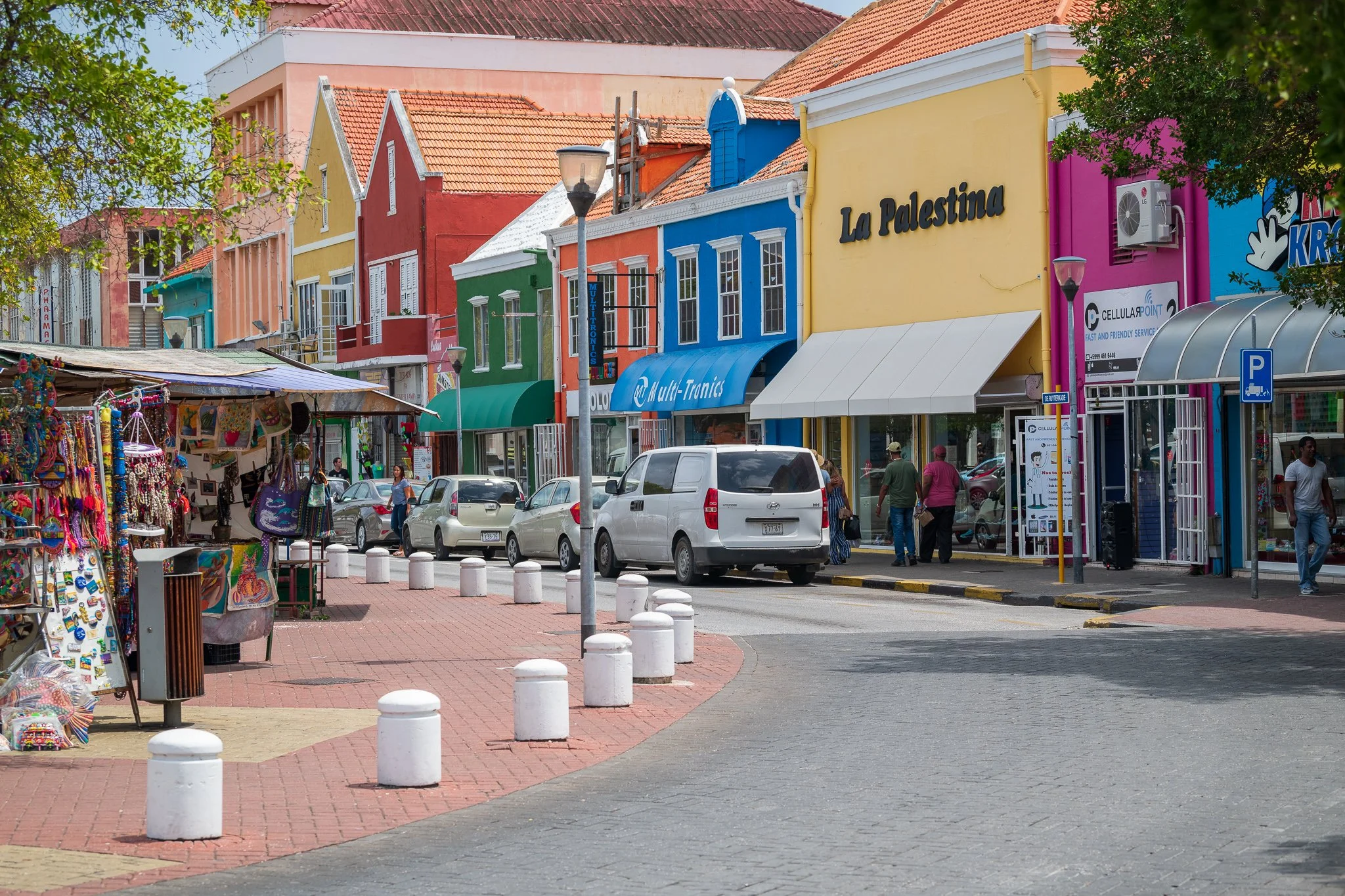 Colorful storefronts on a city street with parked cars and pedestrians, selling souvenirs and clothing.