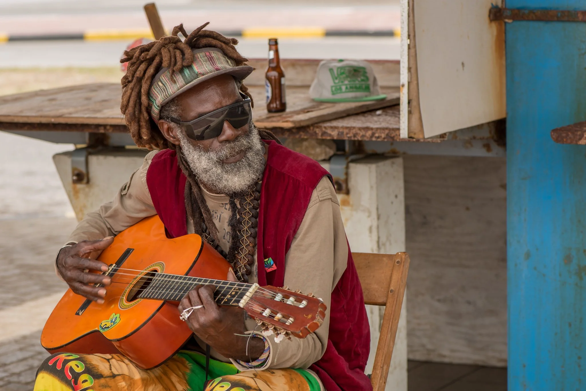 A man with gray dreadlocks wearing sunglasses, a beige shirt, a red vest, and colorful pants, playing an orange acoustic guitar while sitting on a wooden chair near a rustic stand with bottles and bags.