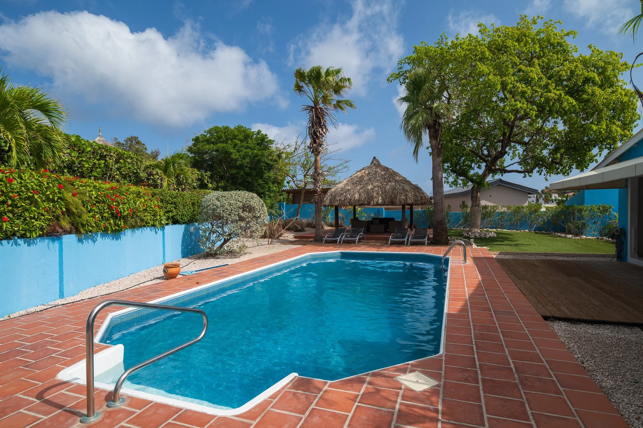 Residential backyard with swimming pool, brick patio, lush green trees, and a thatched-roof gazebo with lounge chairs underneath.