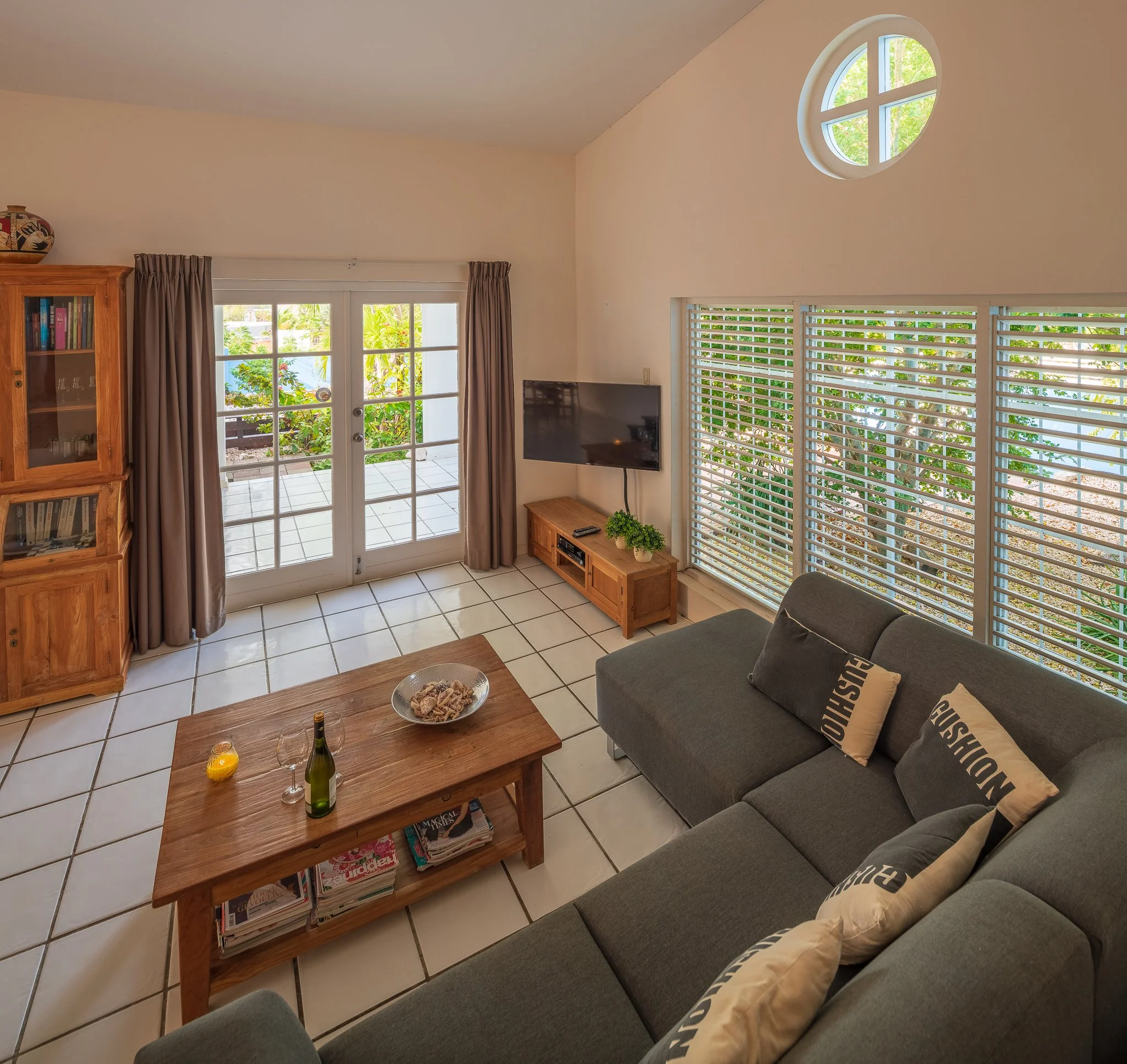 Living room with glass sliding doors leading to patio, a sectional sofa with decorative pillows, a wooden coffee table with magazines, a wall-mounted TV, a wooden entertainment stand with plants, and windows with blinds.