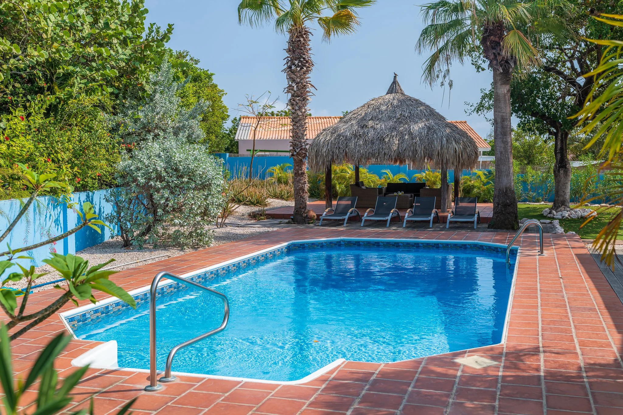 A swimming pool surrounded by a red brick deck, tropical trees, and a thatched roof tiki hut with lounge chairs underneath.