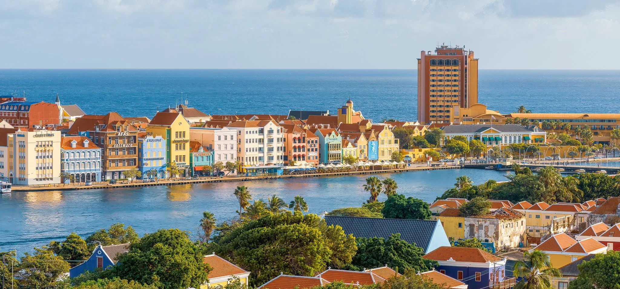 Colorful buildings lining a waterfront with a canal, trees, and a tall hotel overlooking the ocean in the background.
