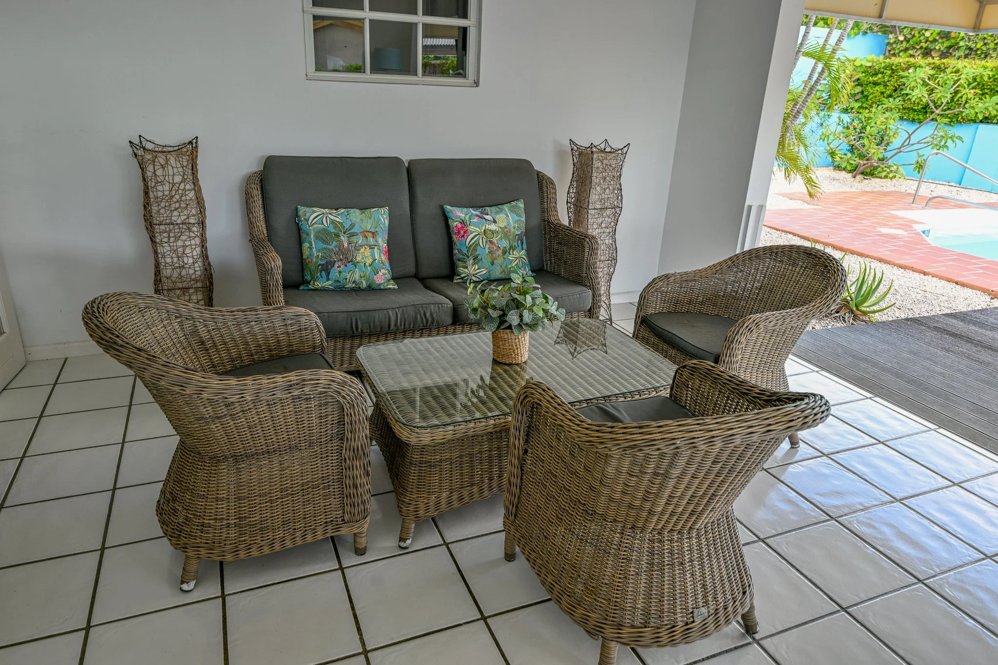Patio seating area with wicker chairs and a glass-top table, positioned near a pool with a pool deck and tropical plants outside.