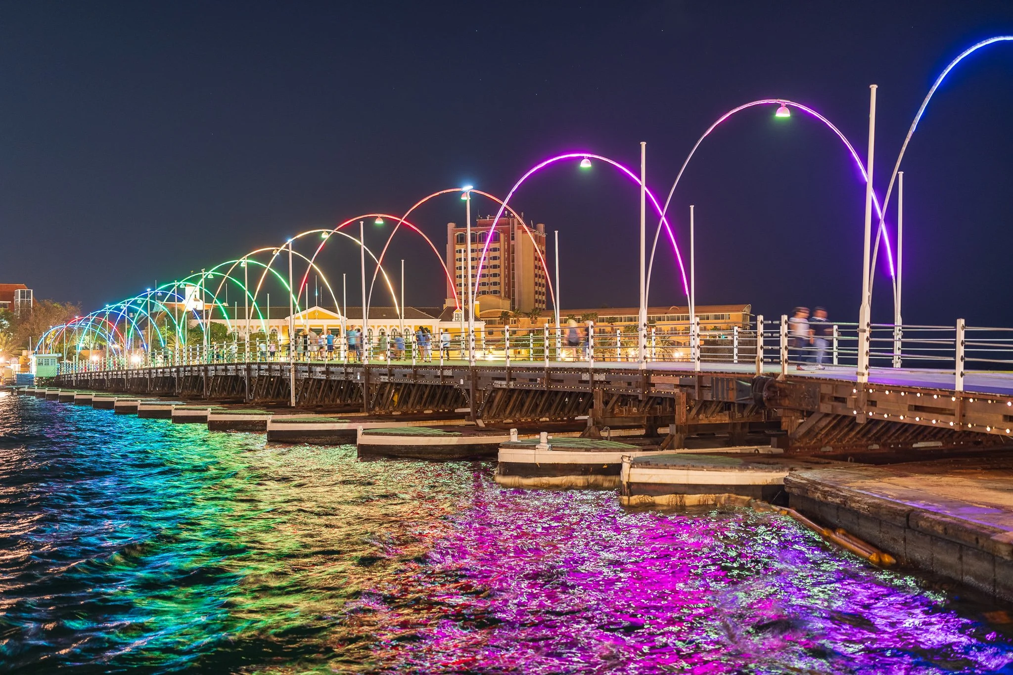 Night view of a pier with colorful rainbow-like lights arching over it, reflecting on the water below, with blurred people walking on the pier and buildings in the background.