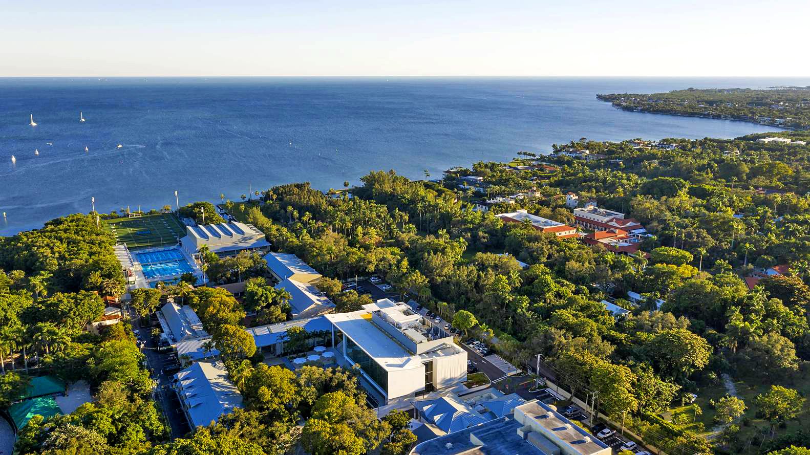 Aerial view of a coastal area with lush greenery, buildings, sports fields, and a large expanse of water with scattered sailboats.