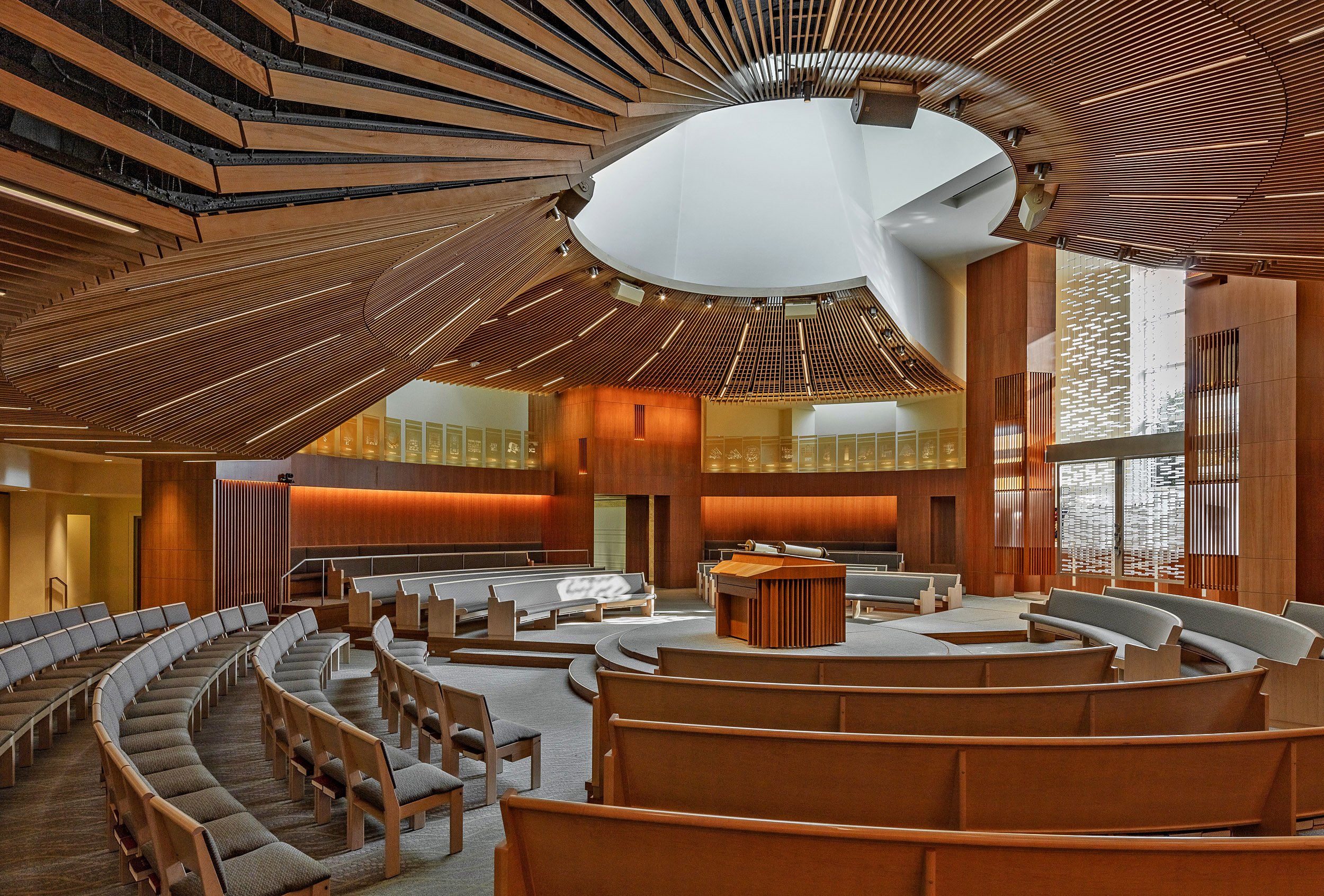 Interior of a modern synagogue with curved wooden benches arranged in a semi-circle, a central pulpit, and a wooden ceiling with a circular skylight.