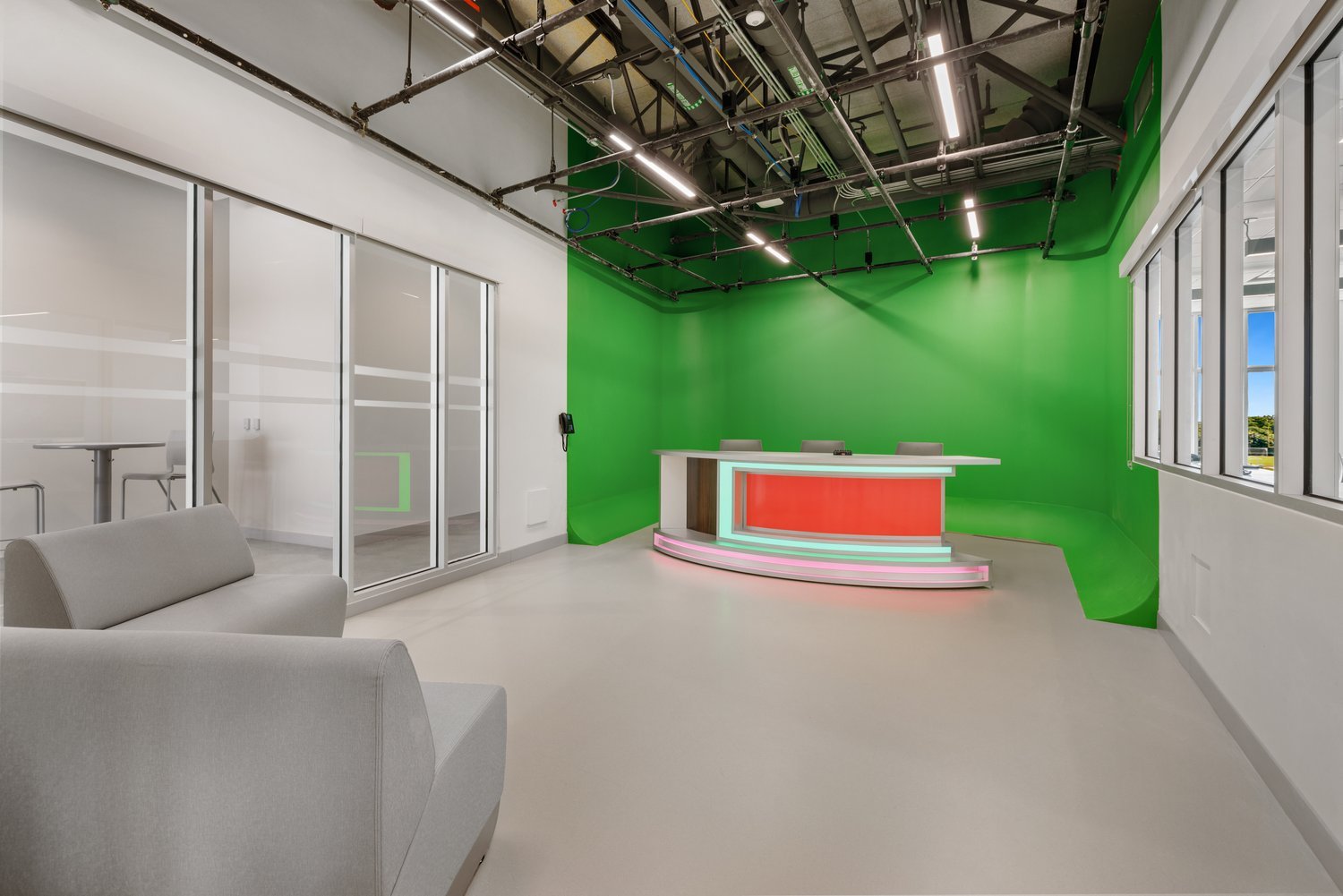 Modern studio with green screen, broadcast desk, gray seating, and glass partition.