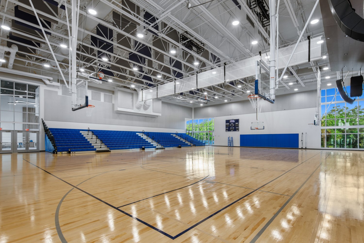 Interior of a modern gymnasium with a basketball court, blue bleachers, and ceiling lighting.