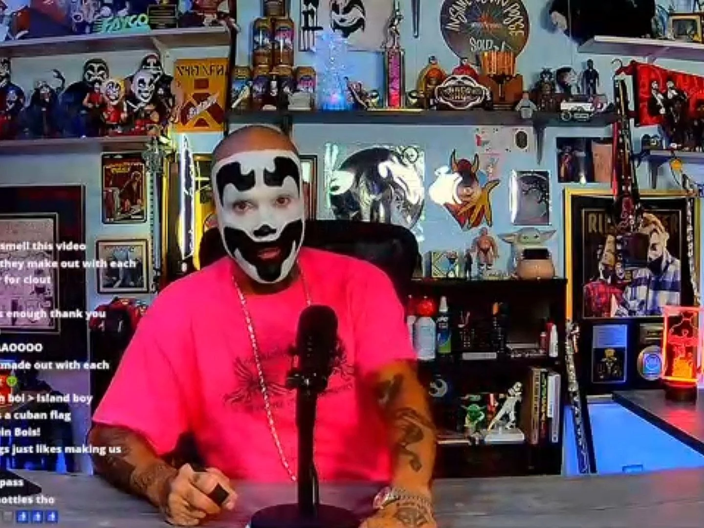 Person with face painted in black and white, wearing a pink shirt, seated in front of a microphone in a room decorated with various posters, artwork, and collectibles, including toys and figures.