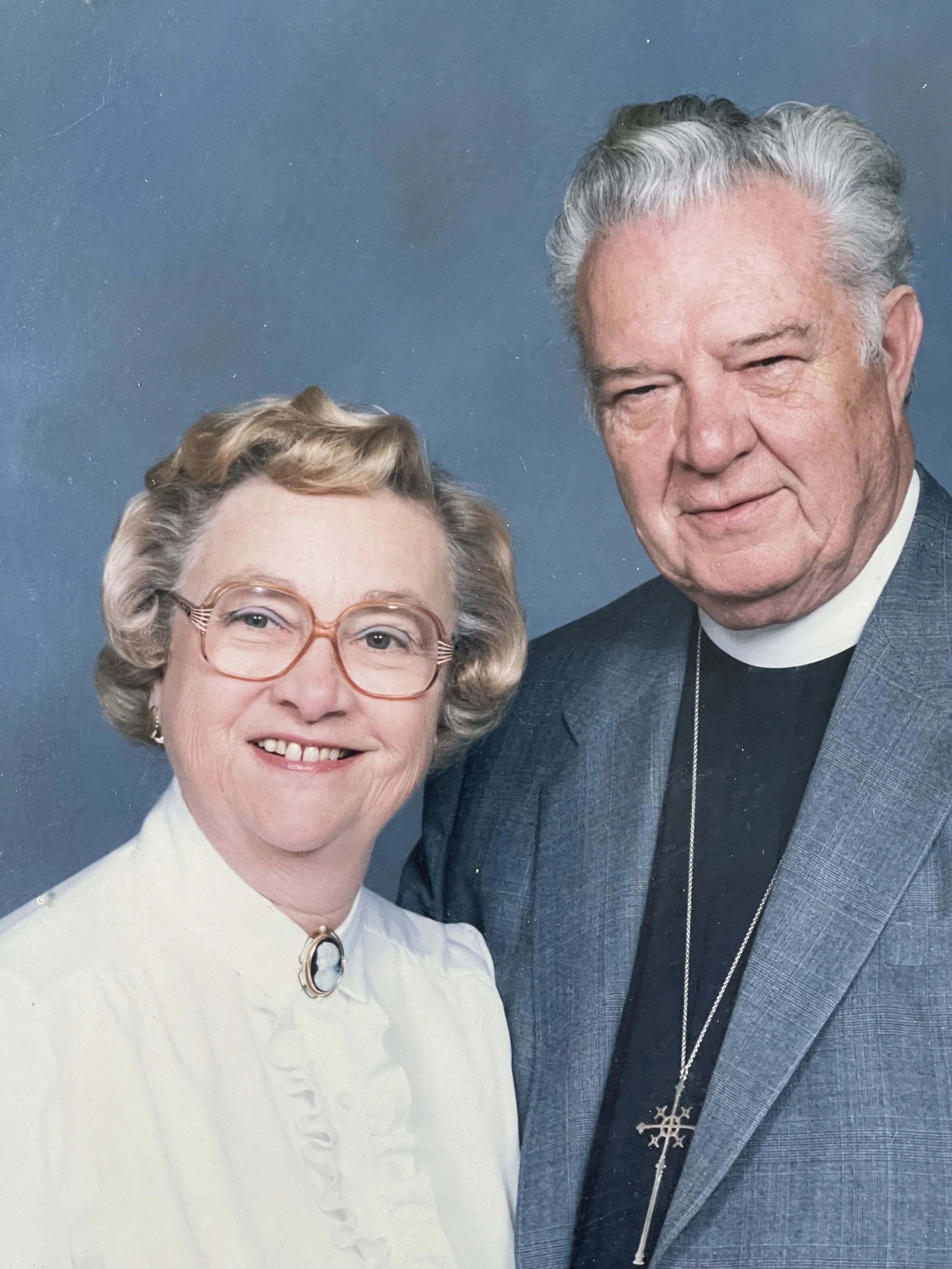 Formal portrait of an older couple against a blue background. The woman wears glasses and a white blouse with a cameo brooch, while the man wears a clerical collar, gray jacket, and cross pendant. Both are facing the camera.