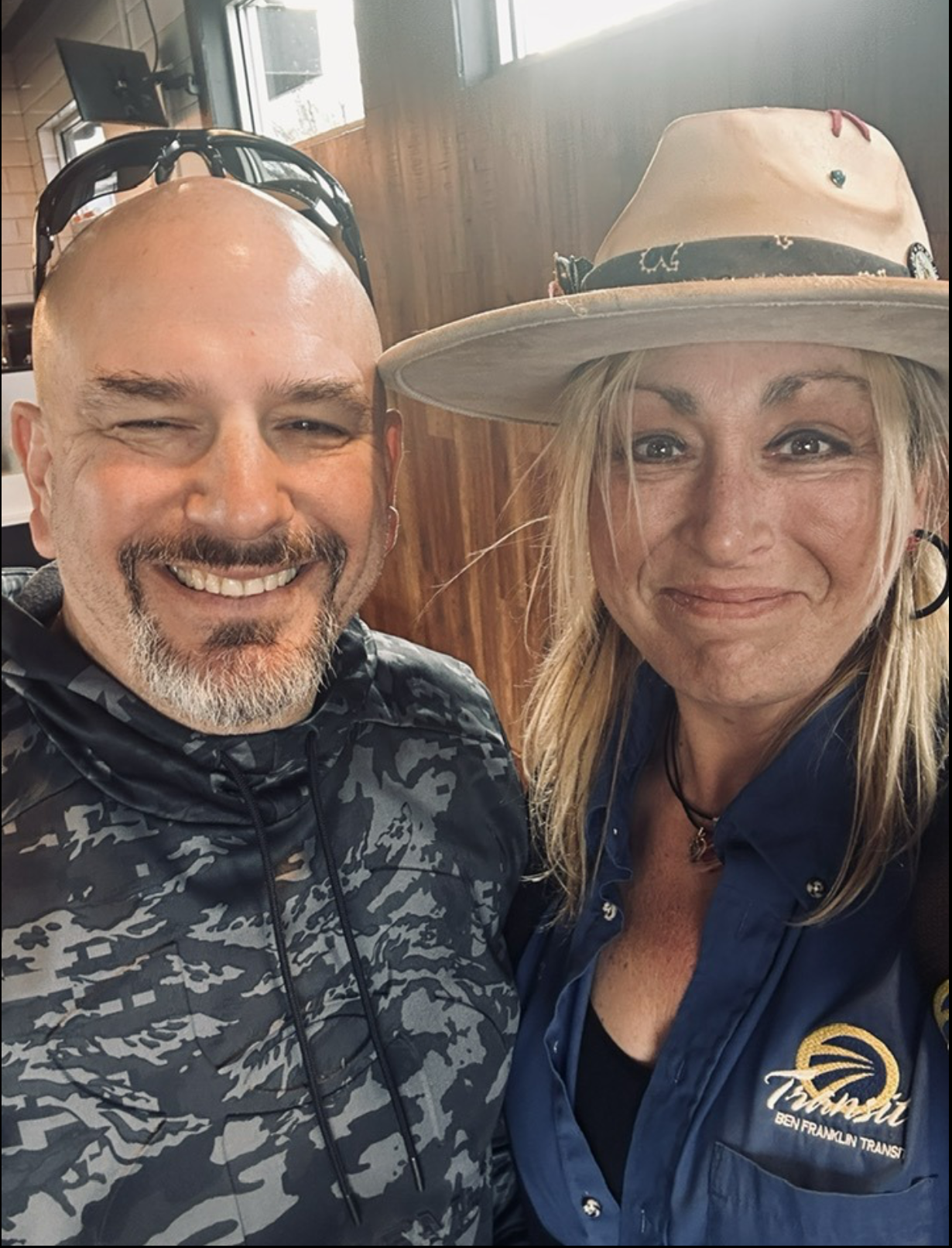 A smiling man with a beard and sunglasses on his head taking a selfie with a smiling woman wearing a wide-brimmed hat, earrings, and a blue shirt with a logo that reads 'Ben Franklin Transit' in a wooden interior setting.