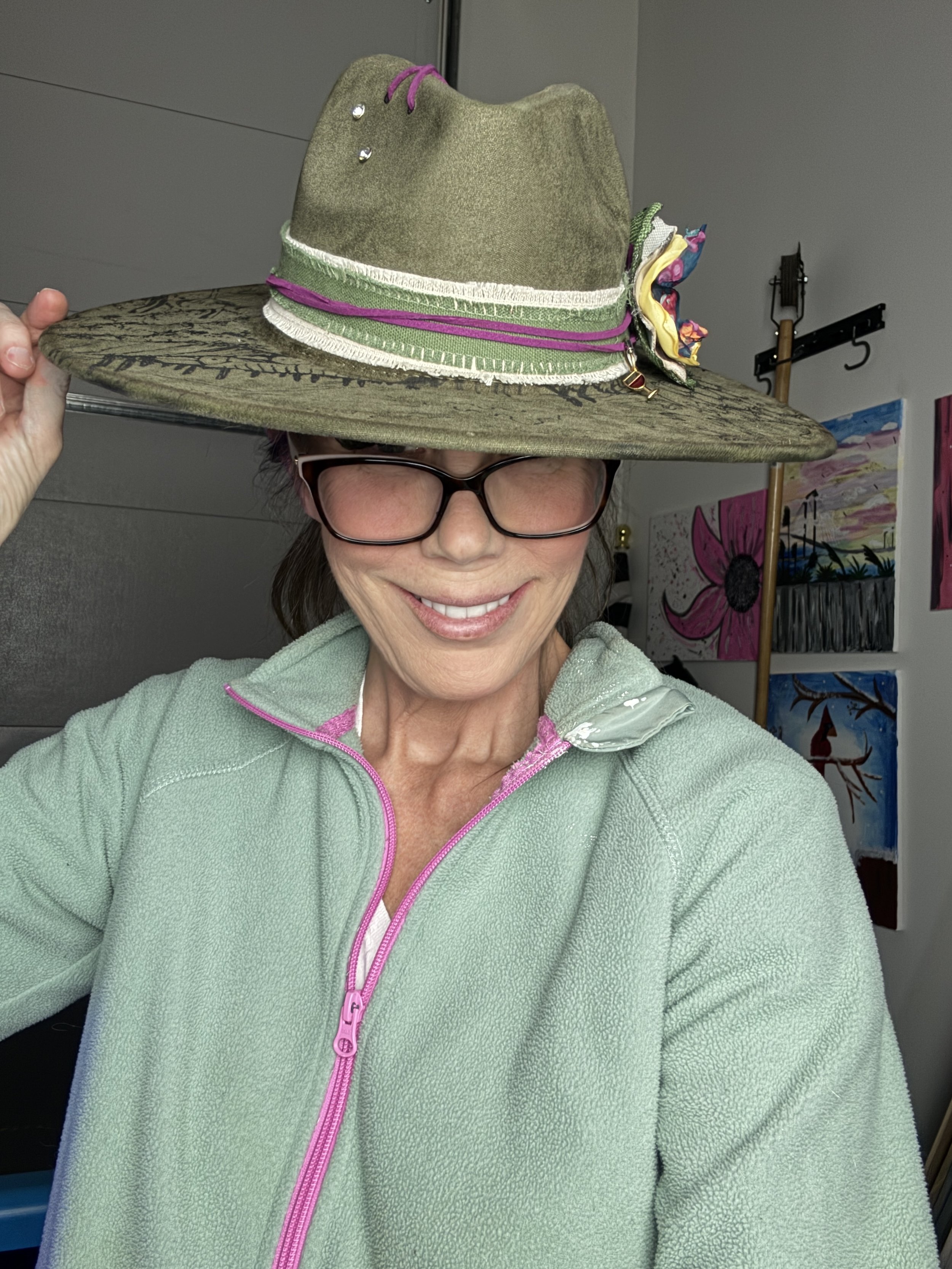 Woman smiling, wearing glasses and a light green fleece jacket, holding a wide brim hat on her head.