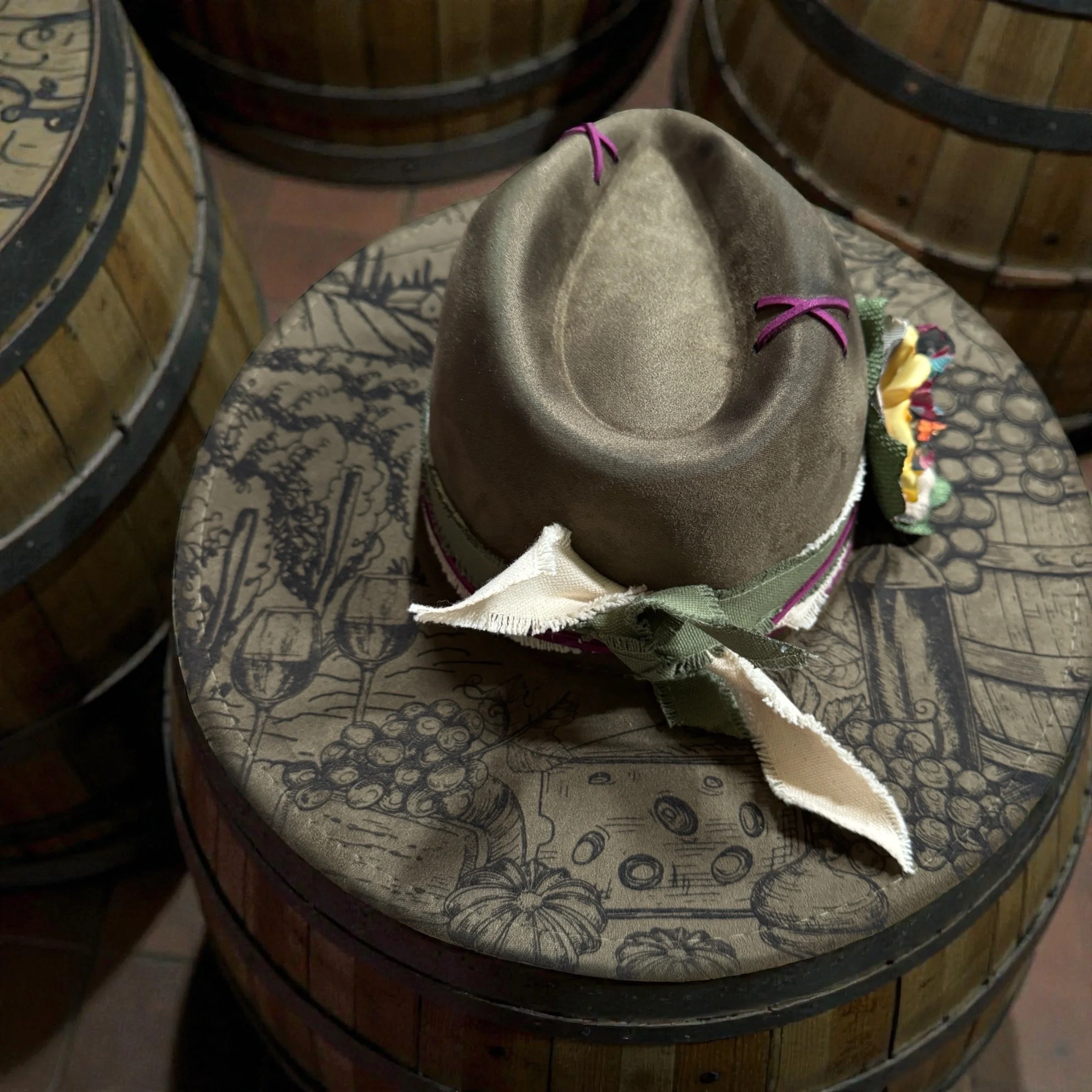 A brown felt hat with purple and green ribbons, placed on a barrel with wood grain and grapevine illustrations.