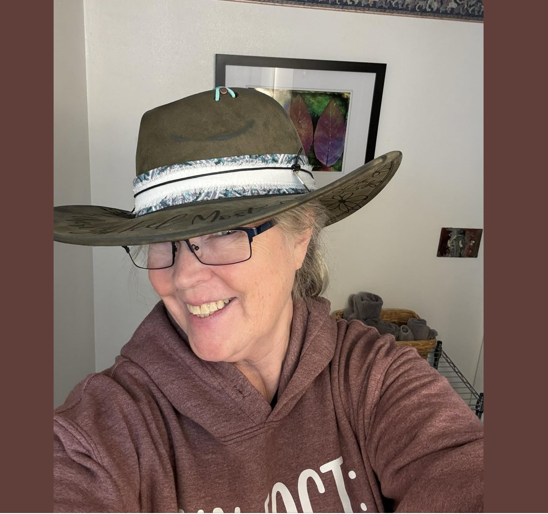A woman smiling and wearing glasses, with a large wide-brimmed hat, in a room with framed artwork on the wall and a basket of rolled towels in the background.