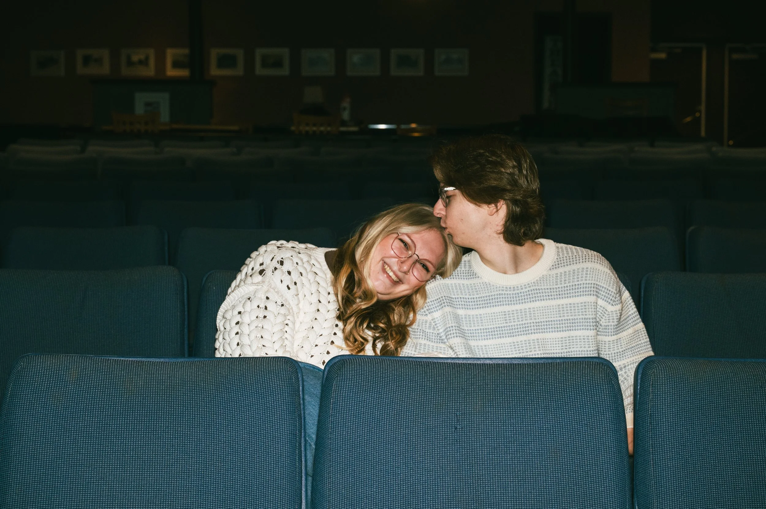 A blonde woman in a large white knit sweater sits in a theatre with a brunette man who is kissing her forehead as she looks off past the camera, smiling.