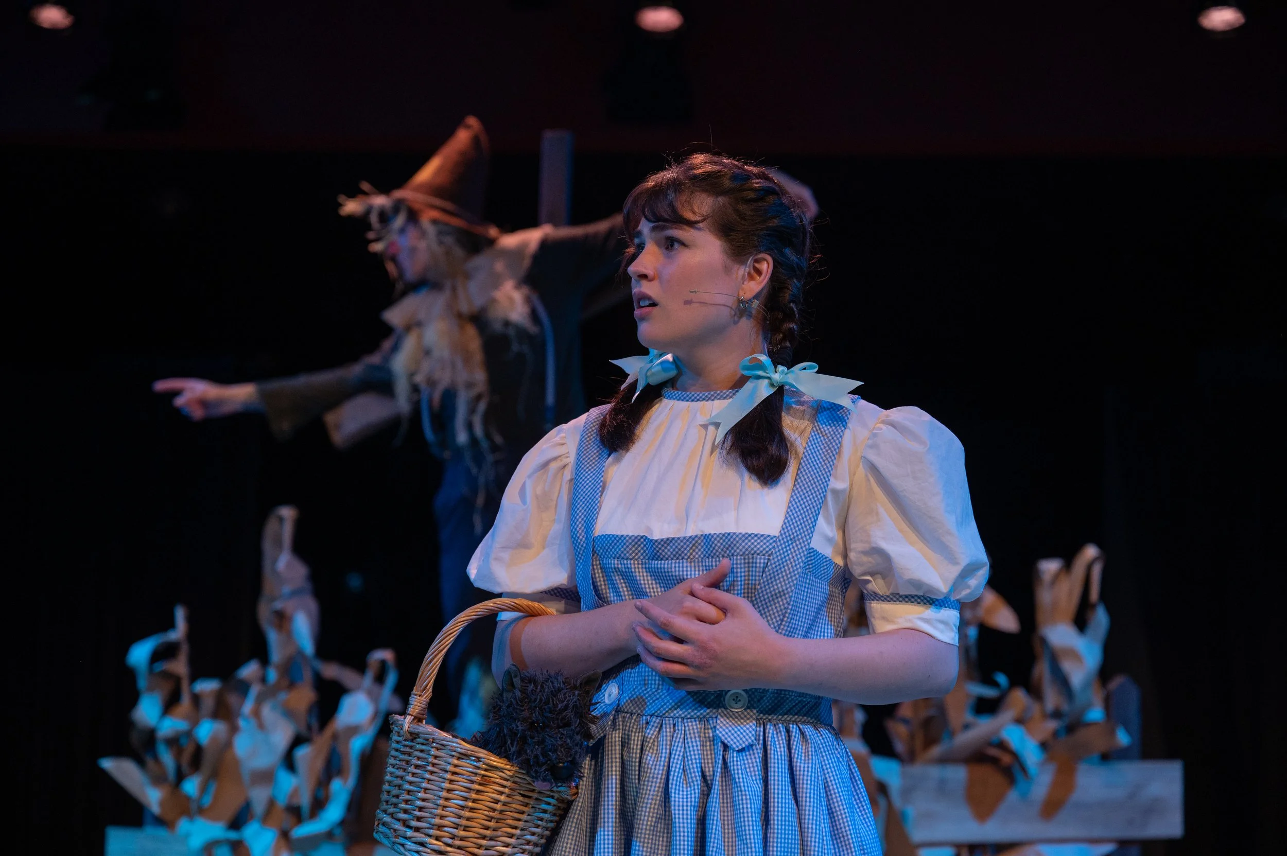 A photo of a girl playing Dorothy in the Wizard of Oz stage production looking to the left while the Scarecrow actor stands in the background pointing to the left. 