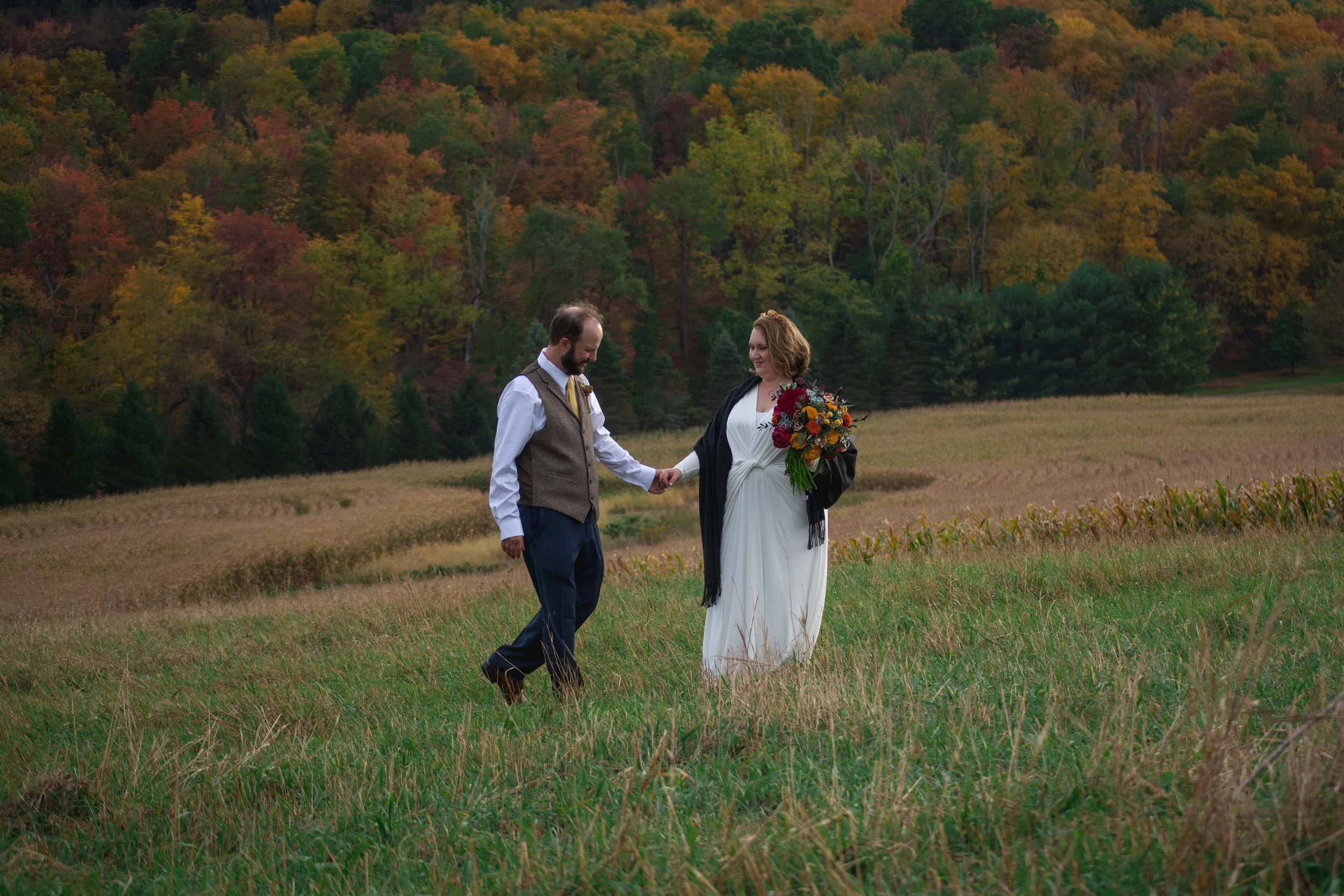 A bride is leading her new husband to her in a field with fall foliage surrounding them in the background.