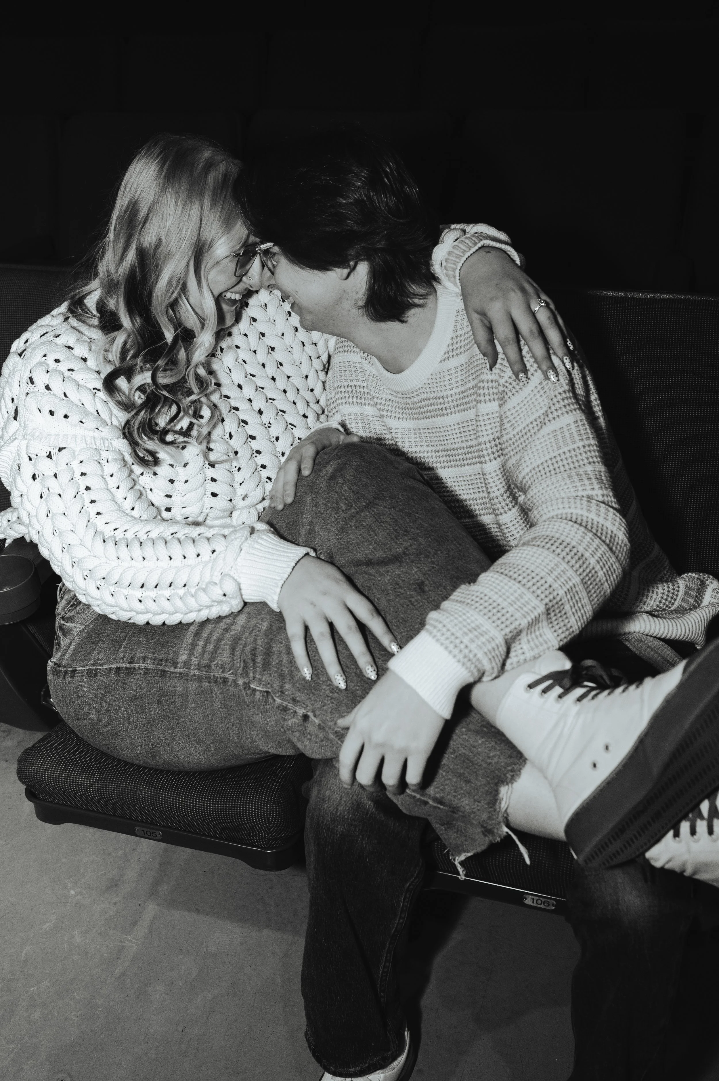 A black and white photo of a man sitting in the front row of a theatre with a woman sitting on his lap as they laugh and touch noses.