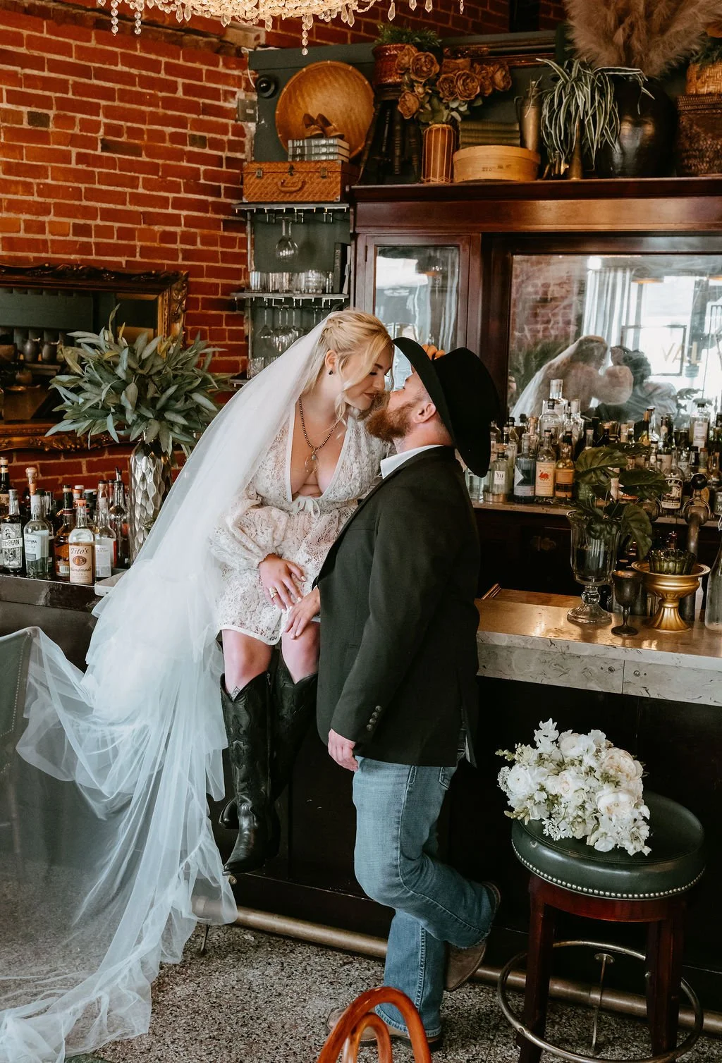 A bride and groom seated on a bar, the bride leans in for a quick kiss.
