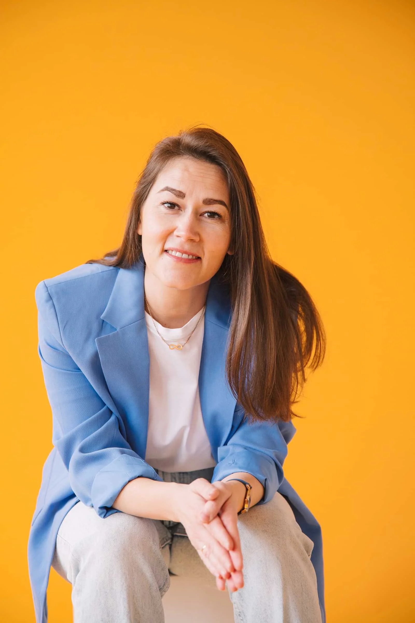 Junge Frau mit braunen Haaren, blauer Jacke, weiße T-Shirt und hellgrauen Hosen sitzend vor gelbem Hintergrund, lächelnd.