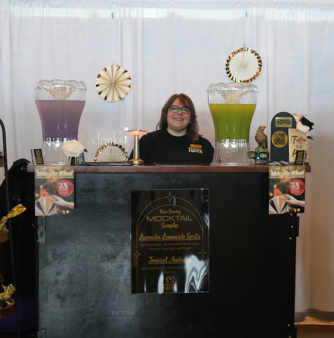 A smiling woman behind a bar counter with two large drink dispensers filled with purple and green beverages, decorated with gold and black accents, and promotional signs for mocktails.