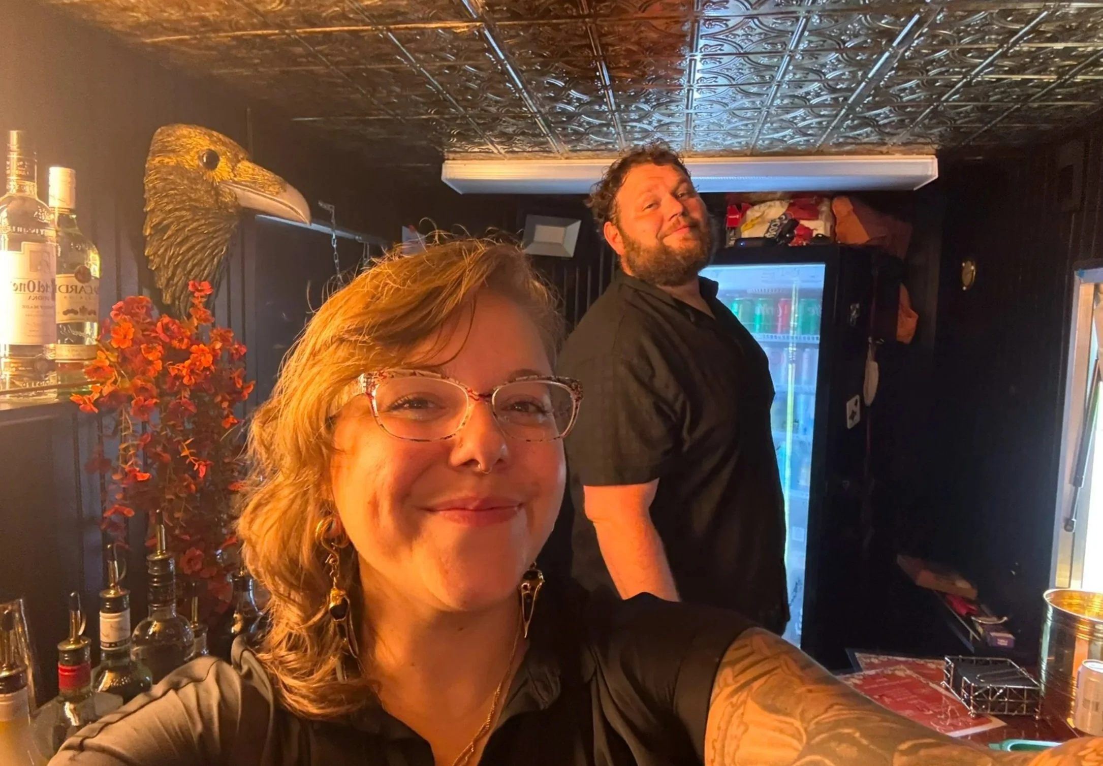 A woman with glasses and earrings taking a selfie with a man in a bar. The woman is smiling, and the man has a smirk. Behind them, bottles of alcohol and a decorative golden raven head are visible. The bar has a dark interior with a tin ceiling.