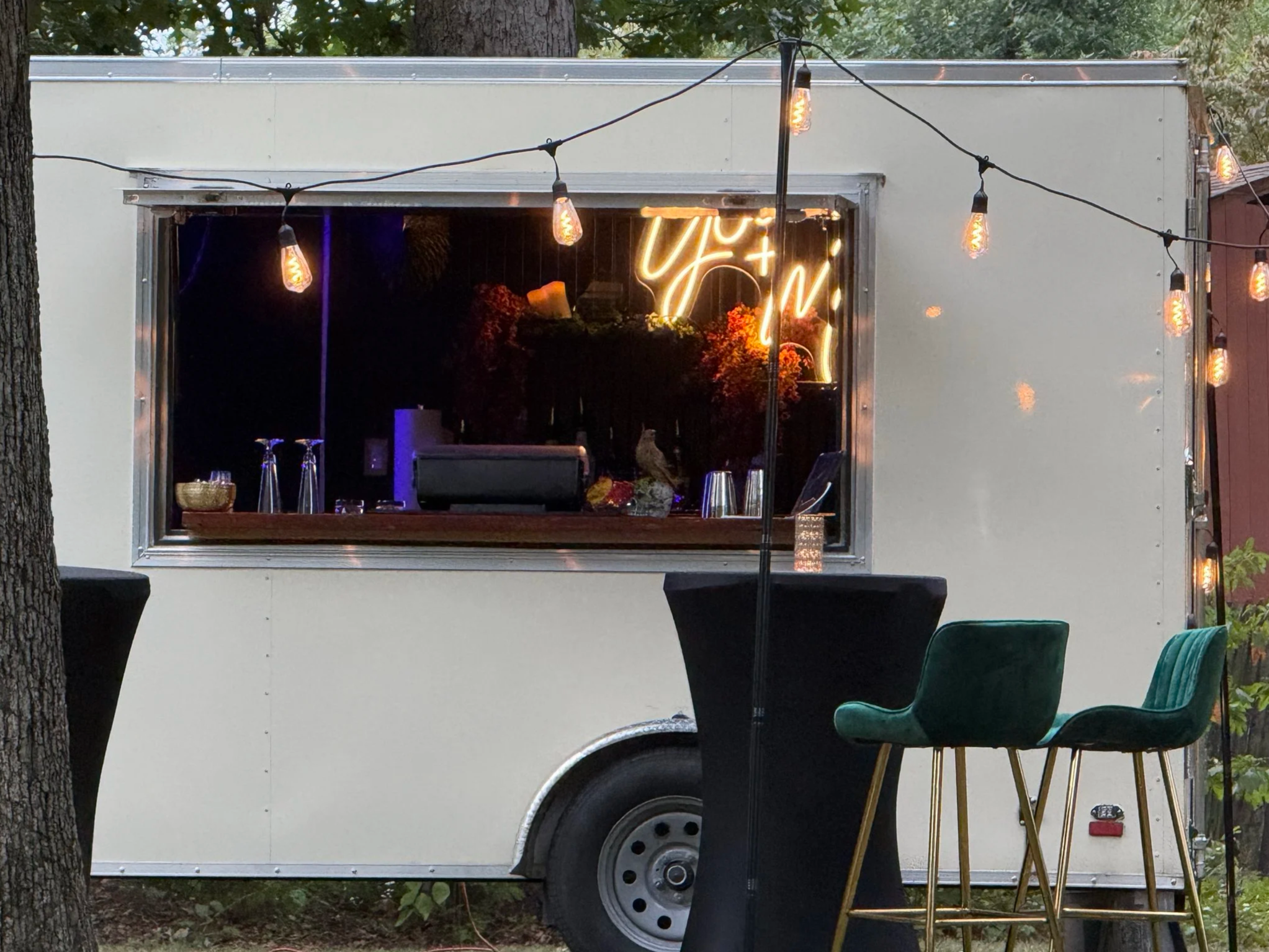 A food truck with a large window serving area, decorated with string lights, parked outdoors among trees. There is a high table with two green chairs outside.