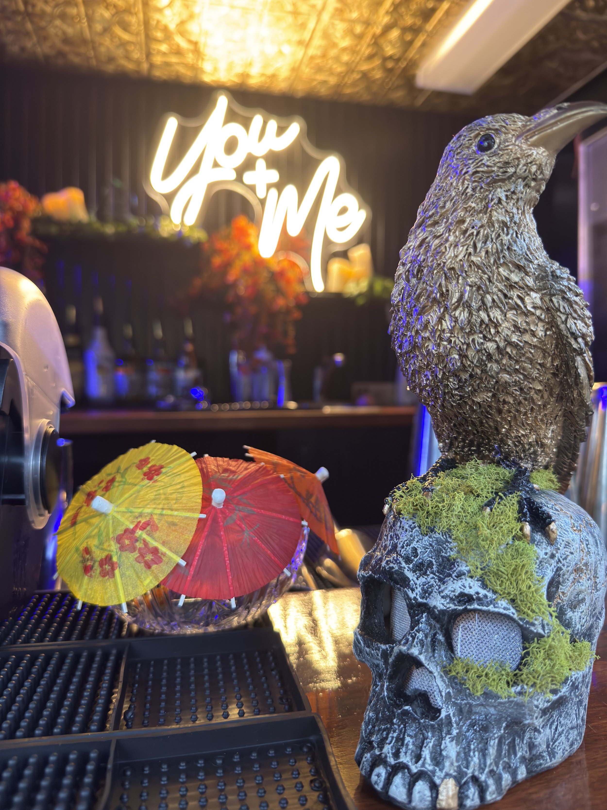Decorative bar with a large metallic owl statue on a skull with green moss, colorful cocktail umbrellas, and a neon sign in the background that says 'You + Me'.