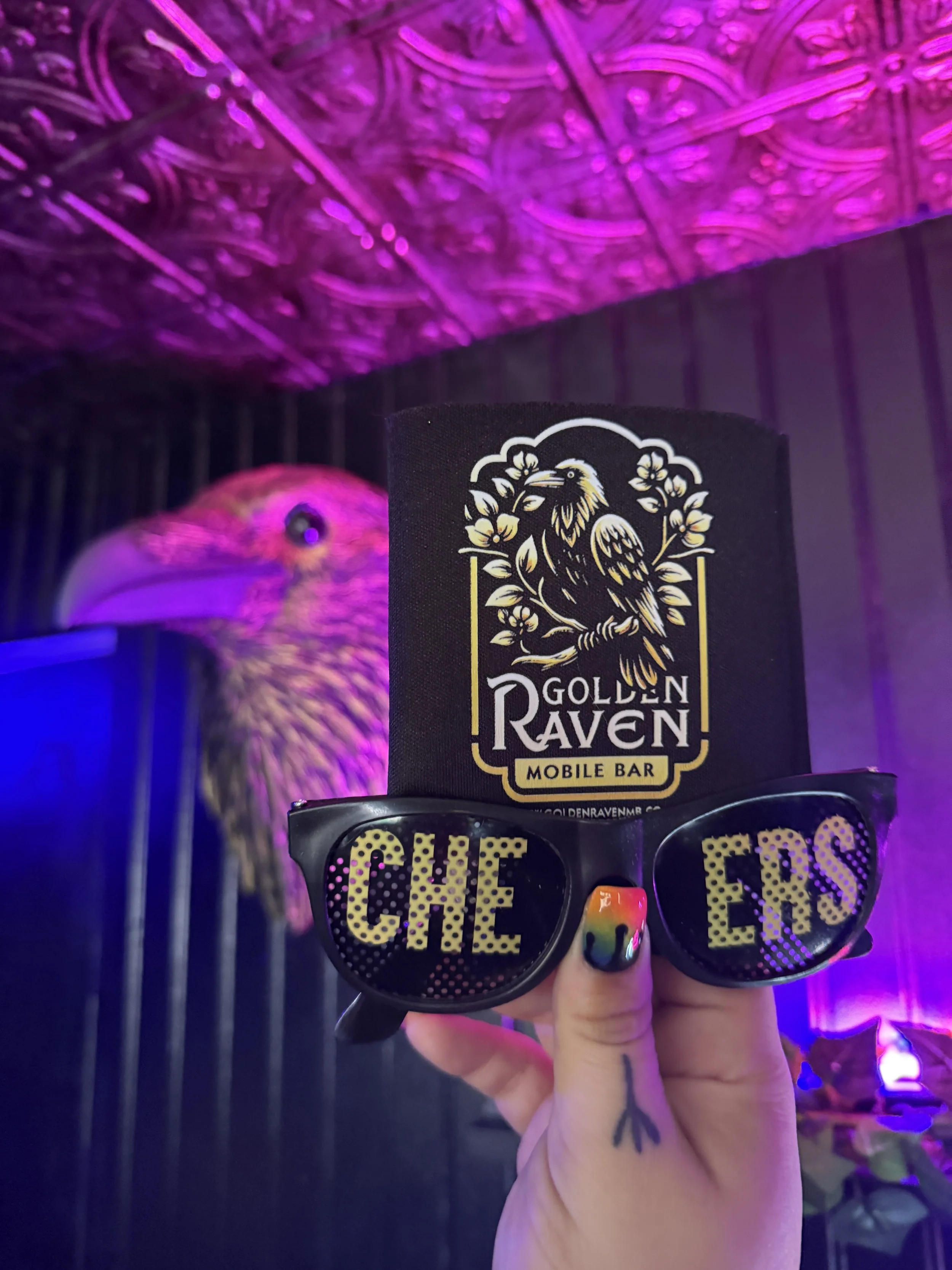 A hand with a small tattoo of an arrow on the thumb holding sunglasses with the words "CHEERS" on the lenses, in front of a black paper cup with a golden raven logo and the words "GOLDEN RAVEN MOBILE BAR". A colorful bird, possibly a parrot, behind a cage is illuminated by purple and pink lighting.