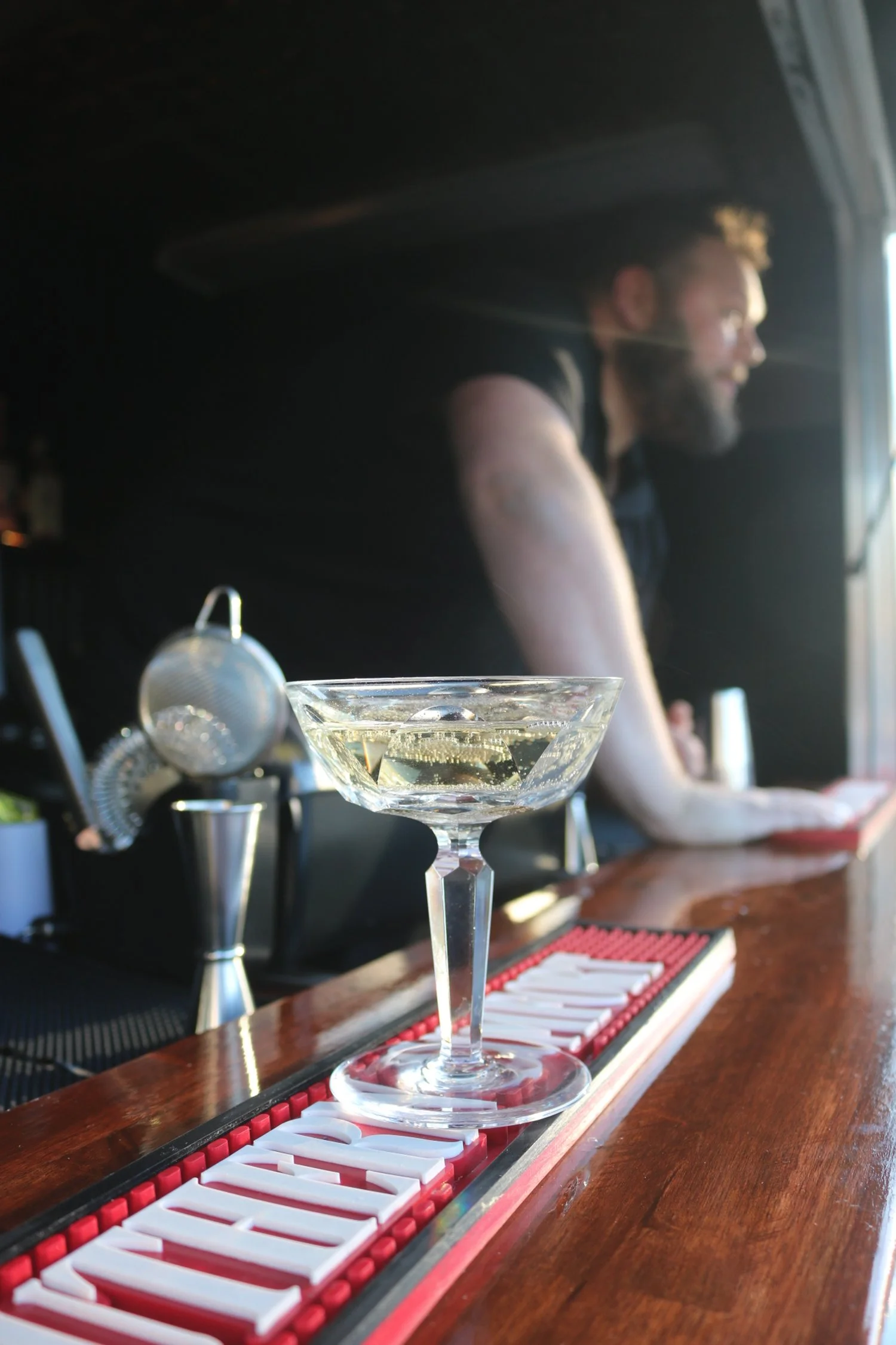 A martini glass filled with a clear cocktail on a bar counter, with a bartender in the background and bar tools nearby.